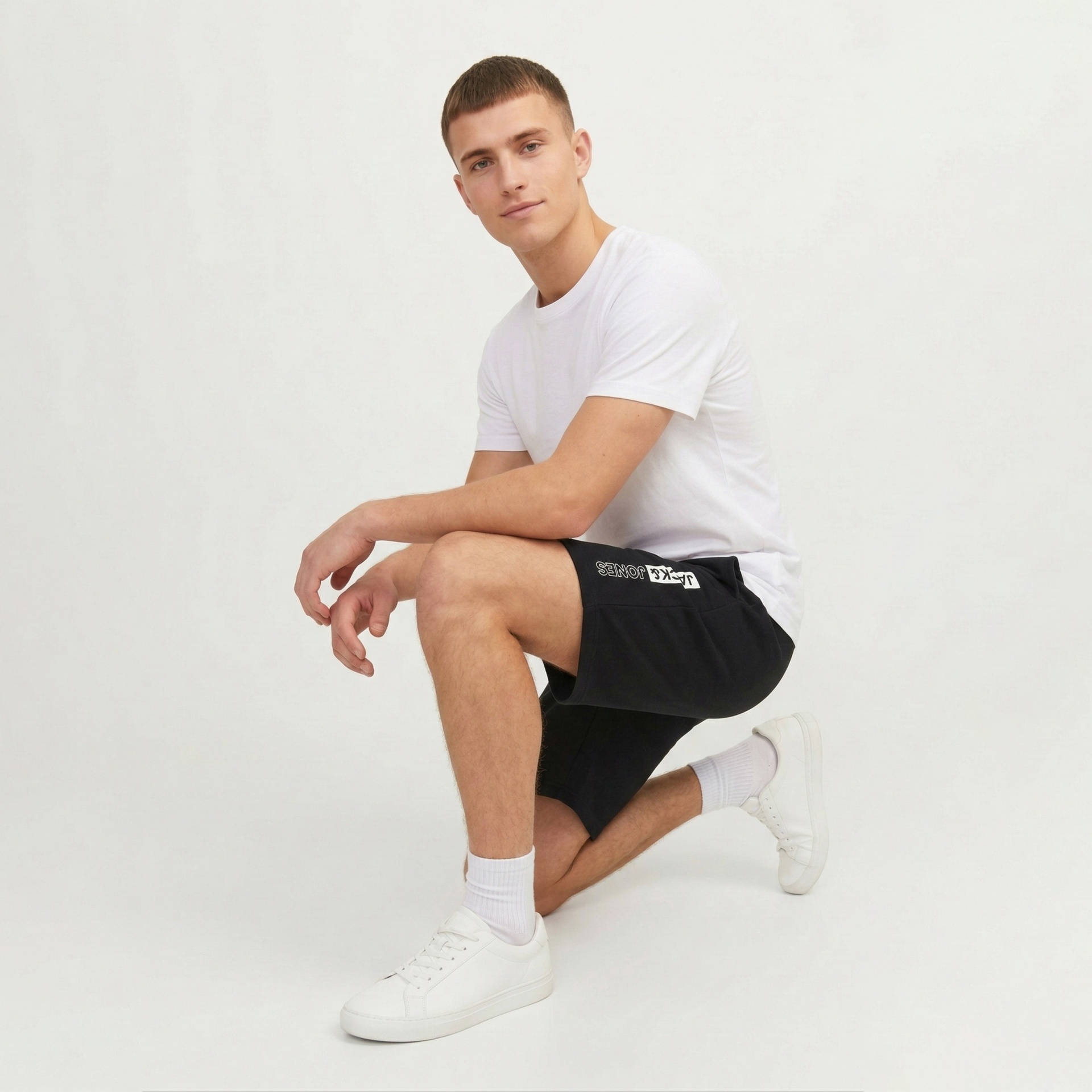 Homem com t-shirt branca, calções pretos com texto JACK & JONES e ténis brancos numa pose ajoelhada