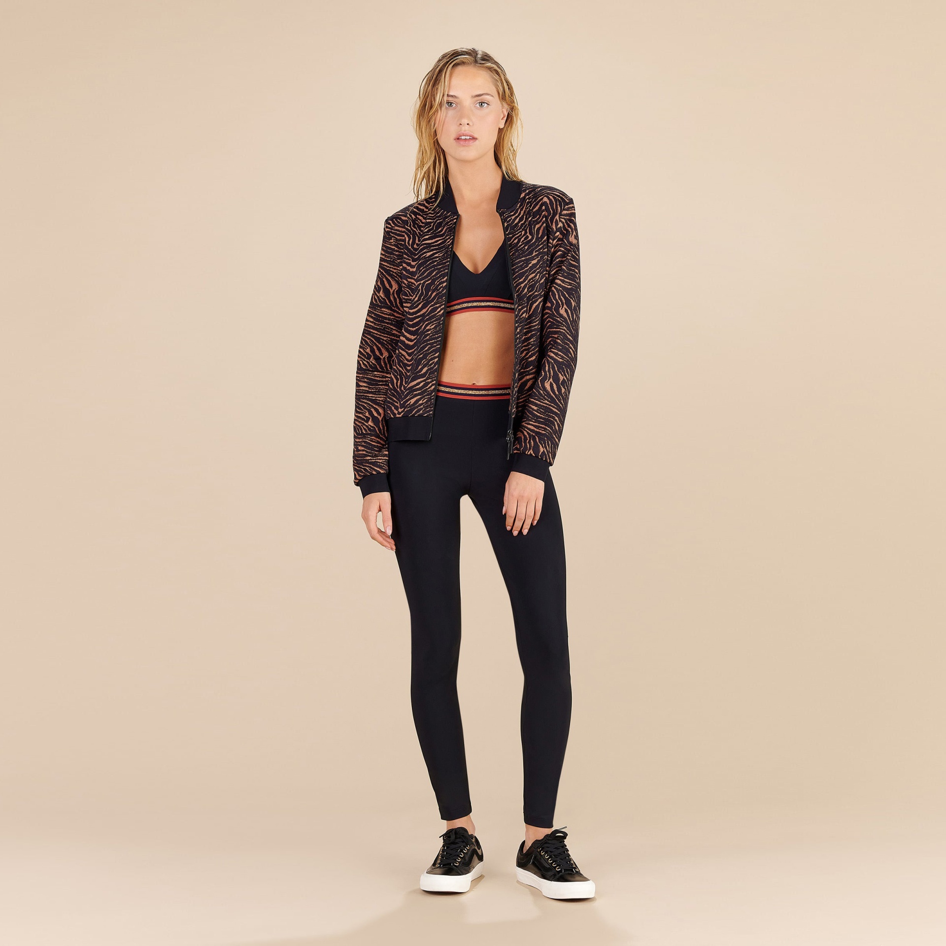 Mulher com casaco bomber estampado, top de desporto e leggings pretas em fundo bege