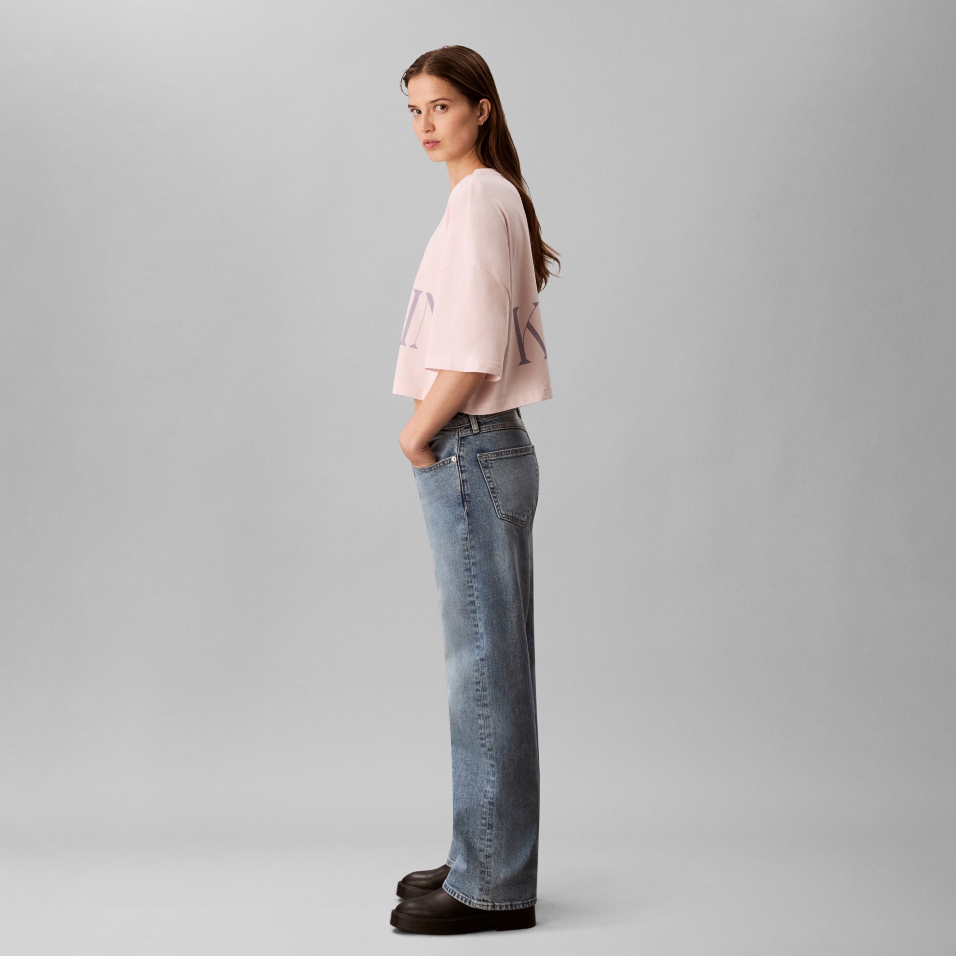 Mulher com camisola rosa de manga curta, jeans azul claro e sapatos pretos em fundo cinza.