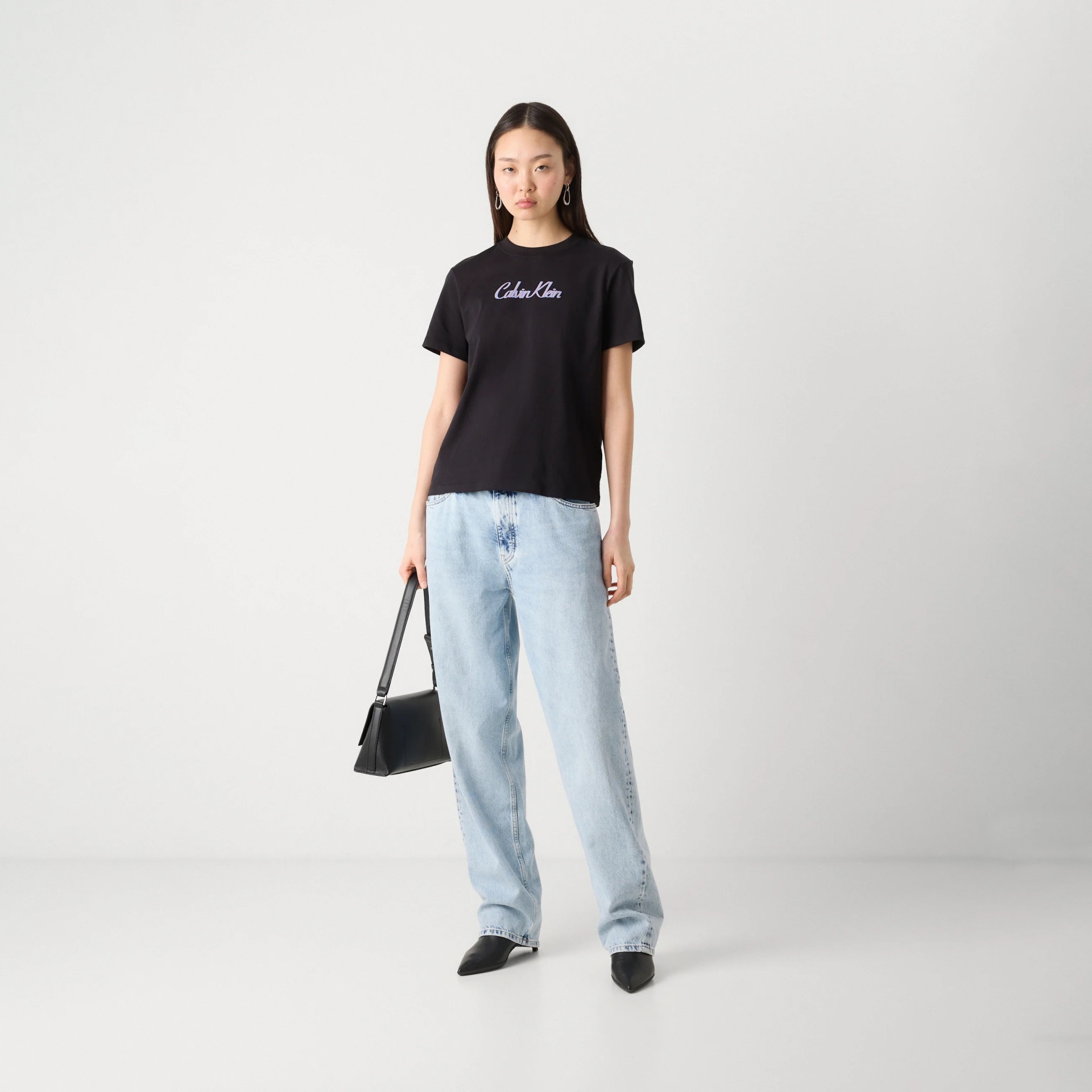 Mulher com t-shirt preta Calvin Klein, calças de ganga azul claro e mala preta, em fundo branco.