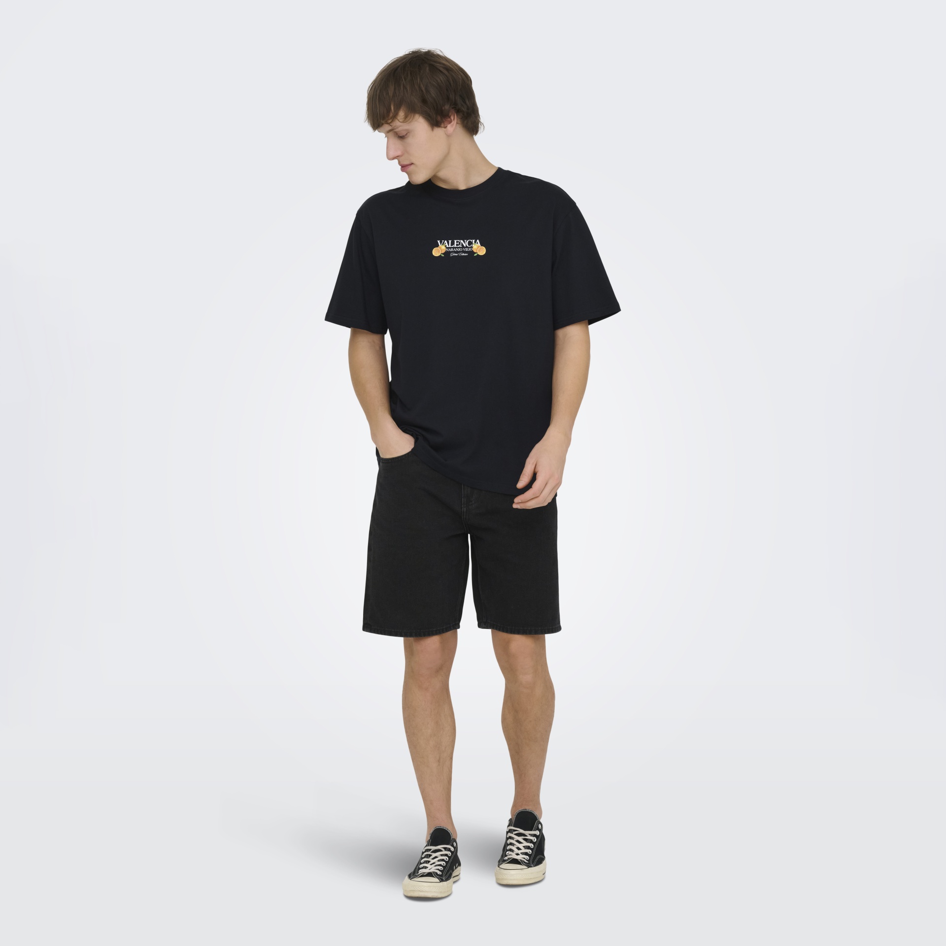 Homem de pé a usar uma t-shirt preta com texto e ilustrações de laranjas, calções pretos e ténis pretos com sola branca.