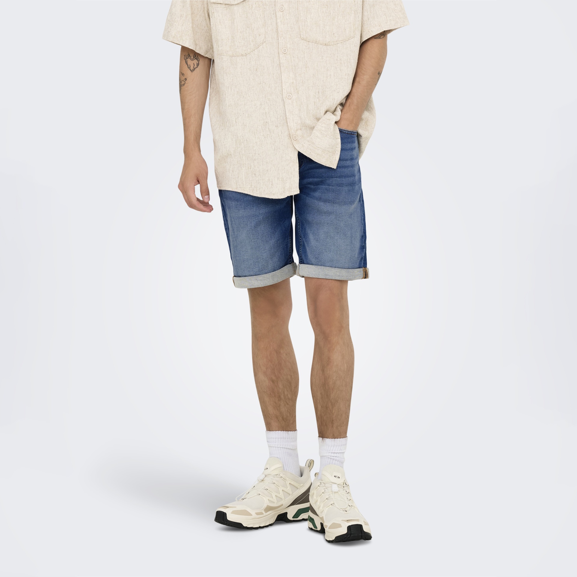 Homem com camisa bege, calções de ganga, meias brancas e sapatilhas desportivas bege.
