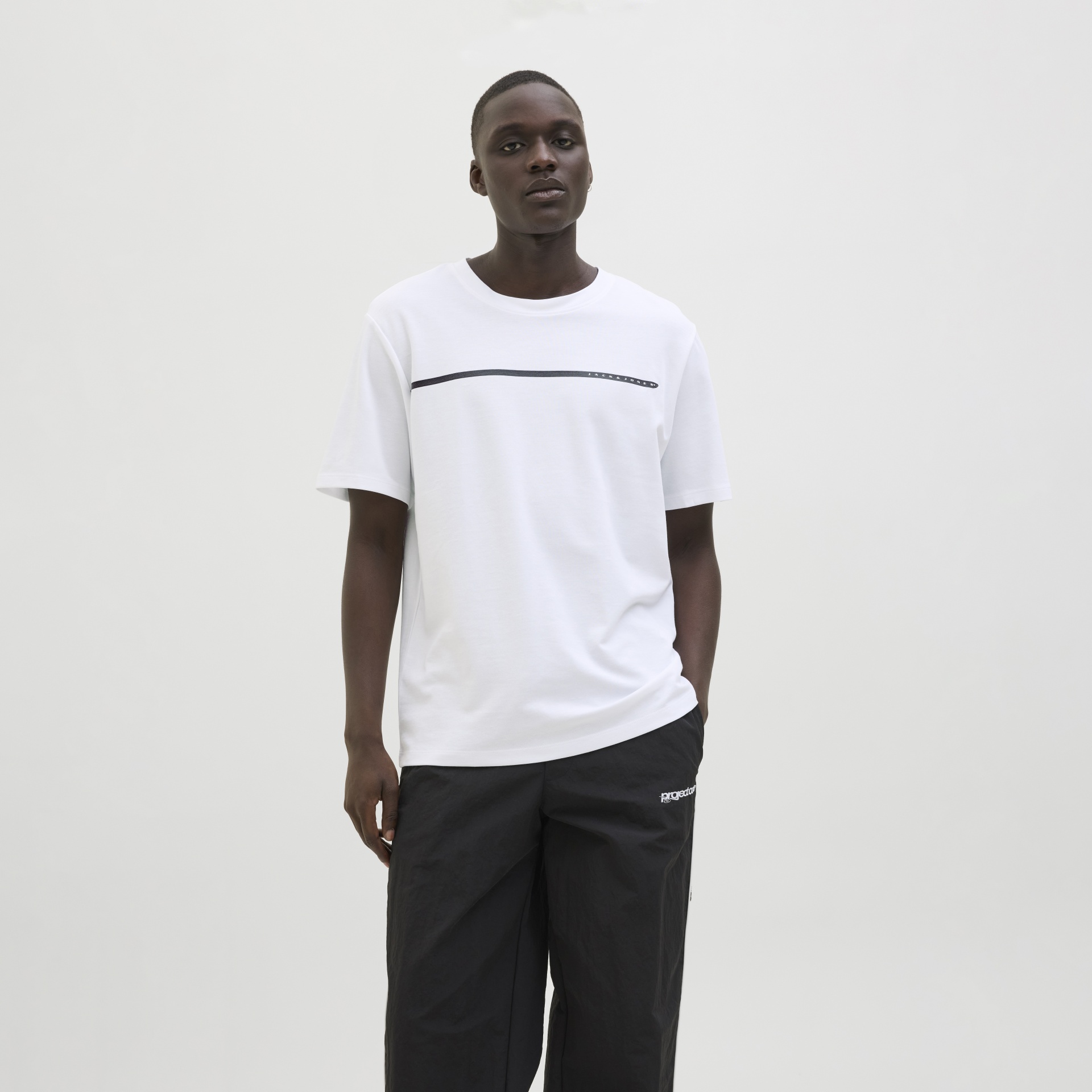 Homem a usar t-shirt branca com linha preta e calças largas pretas em fundo branco