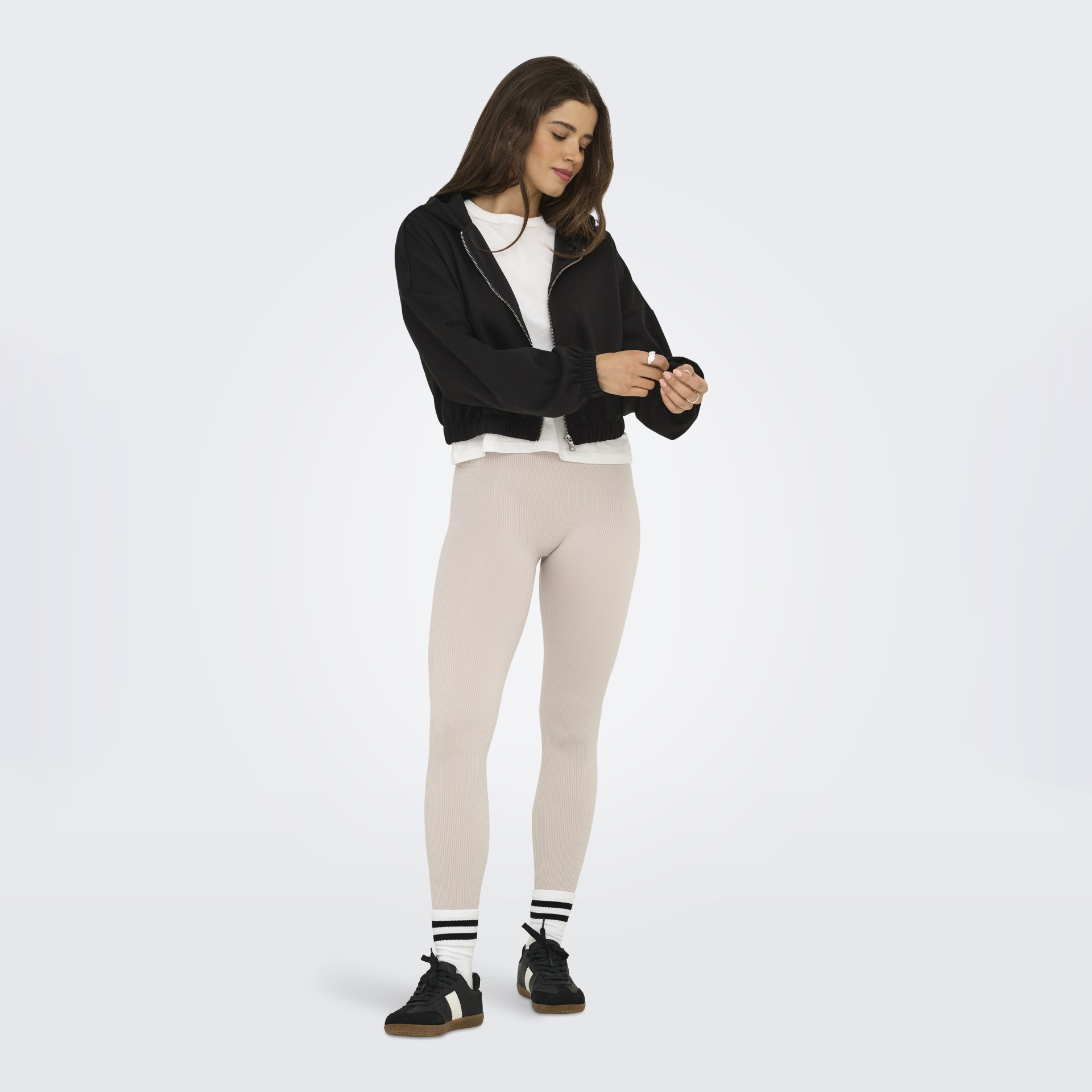 Mulher vestindo casaco preto com capuz fechado, t-shirt branca, leggings bege, meias brancas com riscas pretas e ténis pretos.