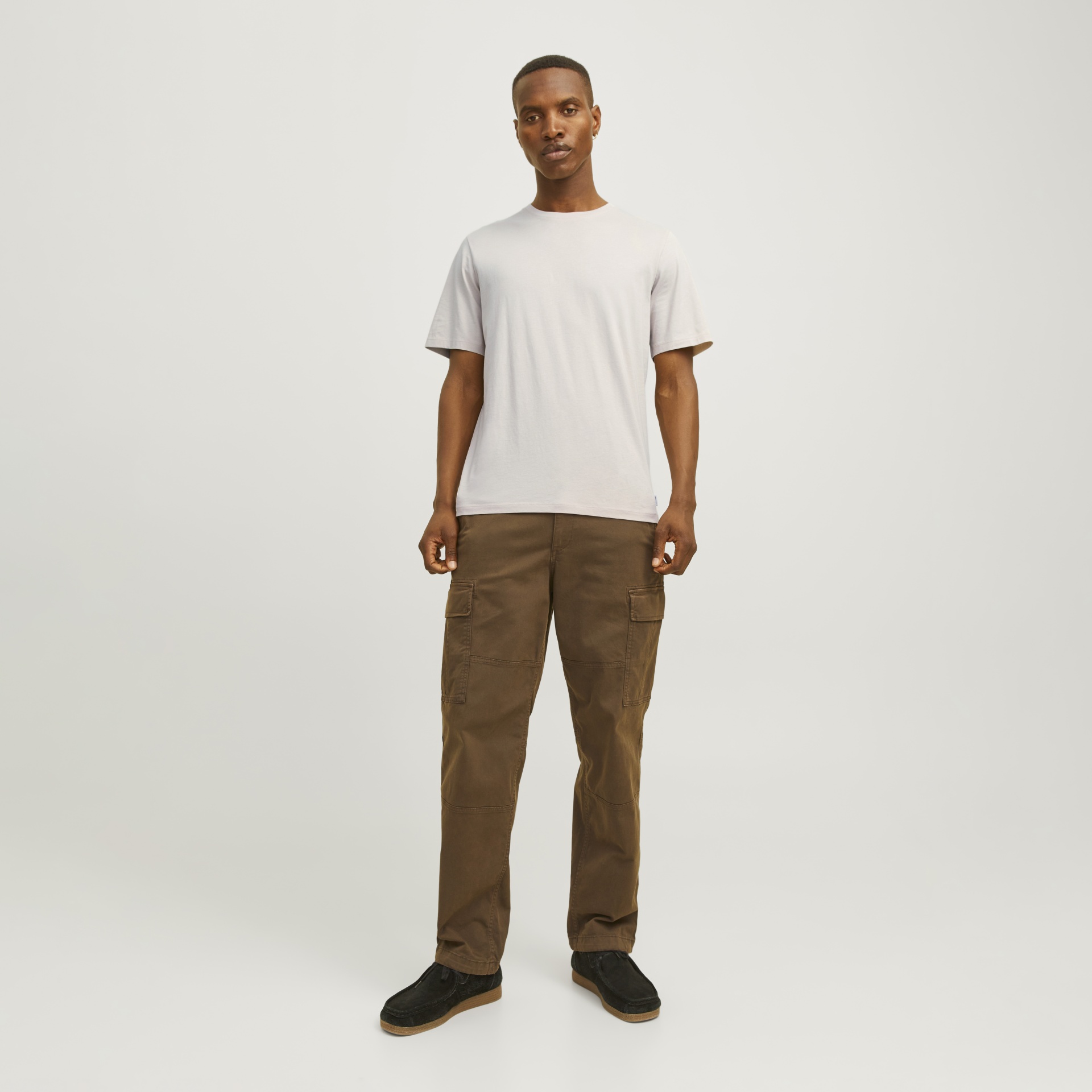 Homem com t-shirt branca, calças cargo castanhas e sapatos pretos em fundo branco.