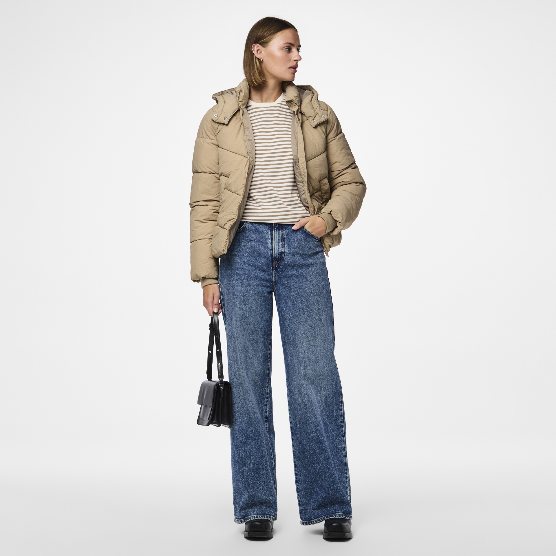 Mulher com casaco acolchoado bege e jeans largos azuis com bolsa preta num fundo branco