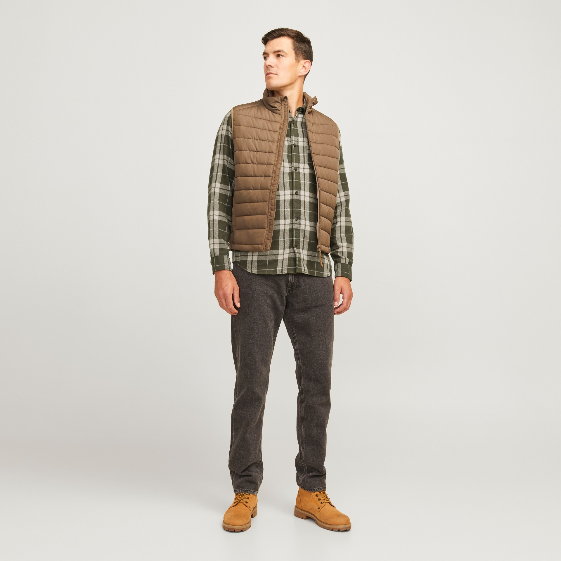 Homem com colete castanho acolchoado, camisa aos quadrados, calças de ganga pretas e botas amarelas