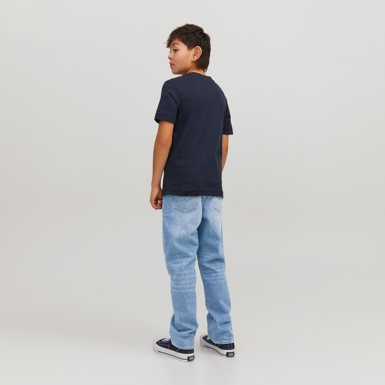 Rapaz de pé vestindo t-shirt azul escura, calças de ganga claras e ténis pretos