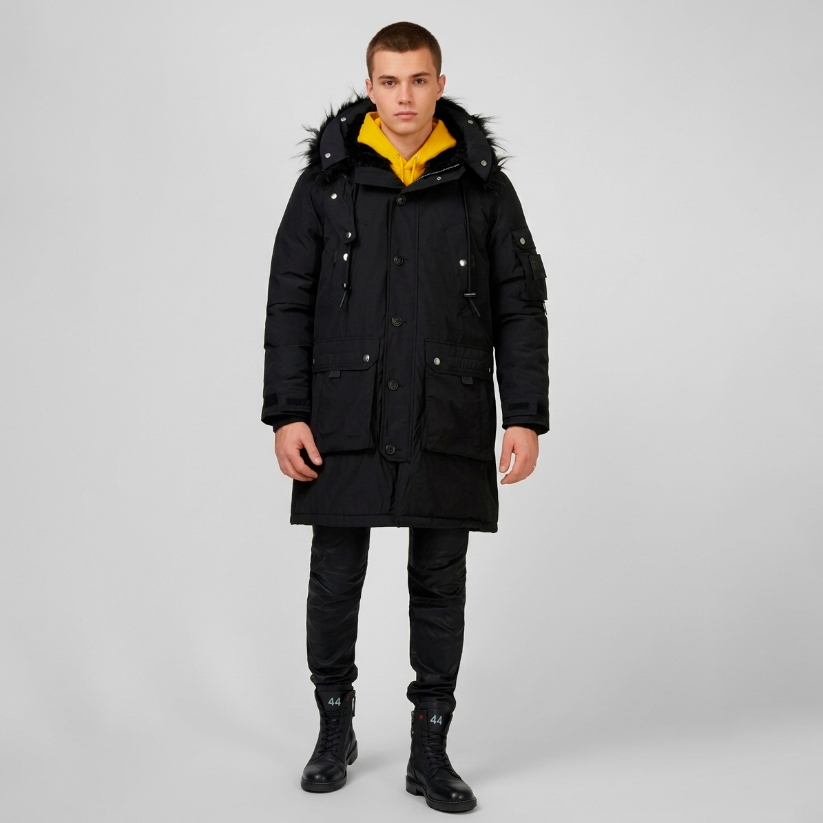 Homem com casaco parka preto com capuz forrado a pele, calças e botas pretas e hoodie amarelo em fundo cinzento