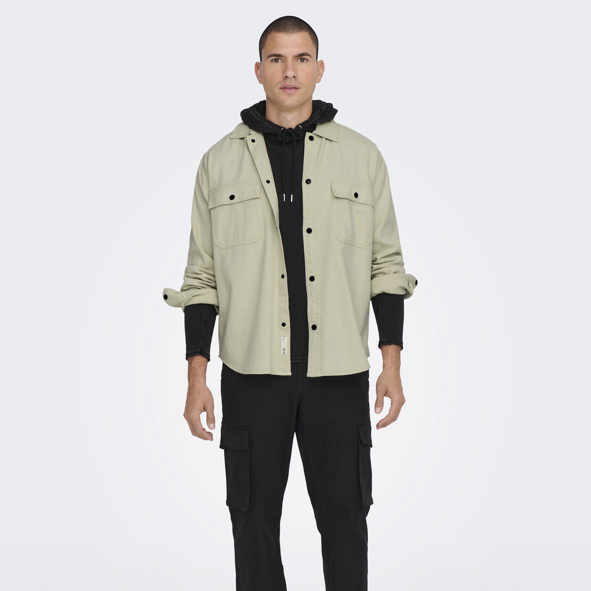 Homem com camisa bege, hoodie preto e calças cargo pretas, fundo branco