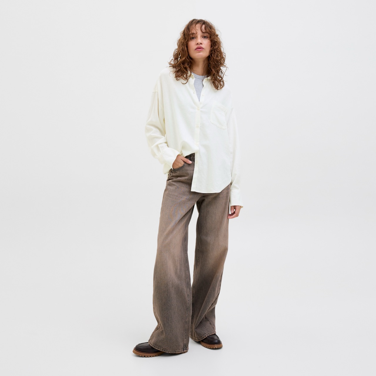 Mulher com camisa branca e calças largas castanhas num fundo branco