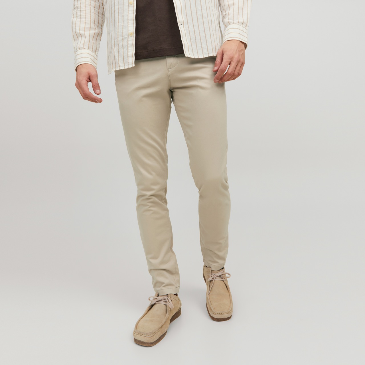 Calças masculinas bege com sapatos castanhos claros e camisa creme às riscas, fundo branco.