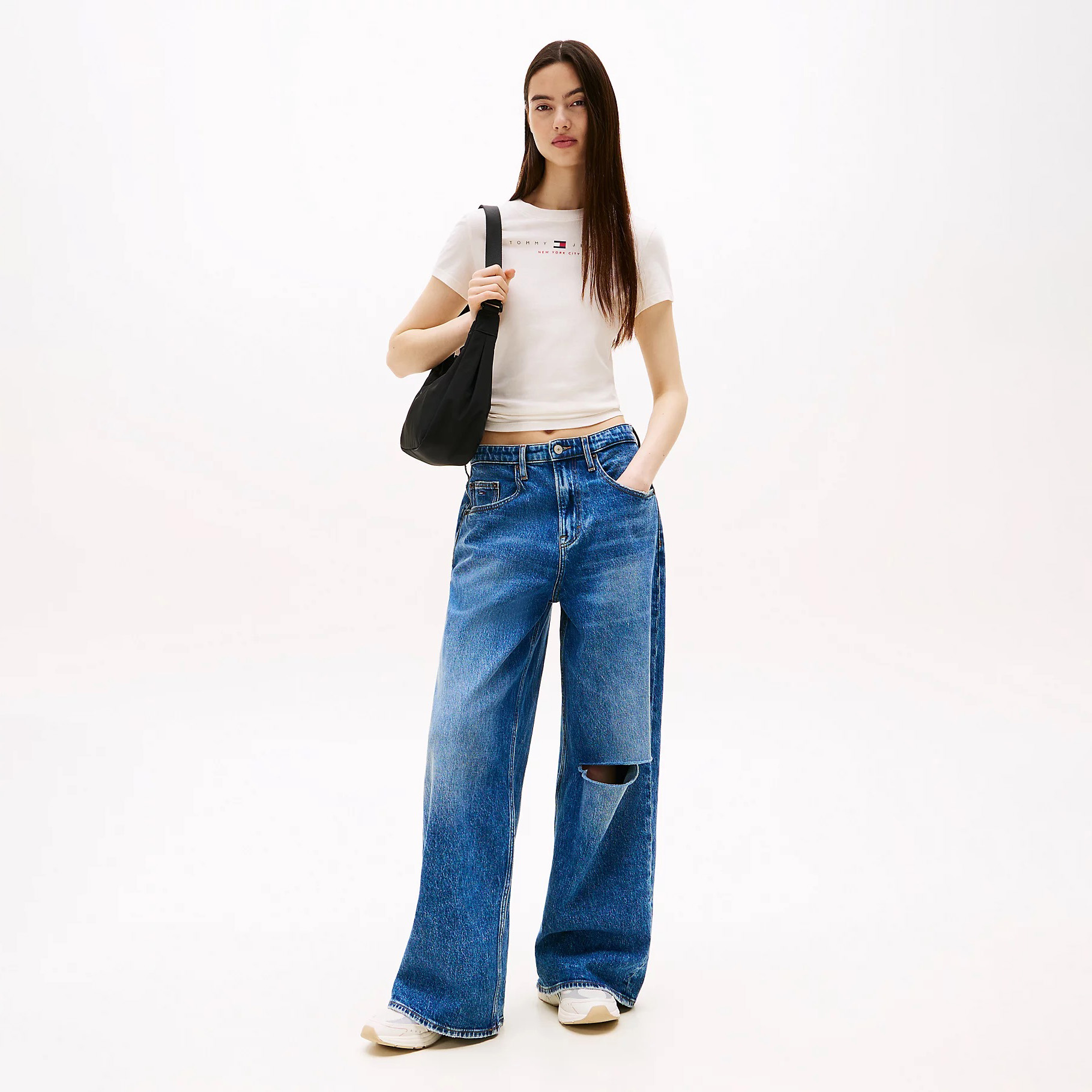 Mulher com t-shirt branca, jeans largos azuis com rasgo, sapatilhas brancas e mala preta ao ombro