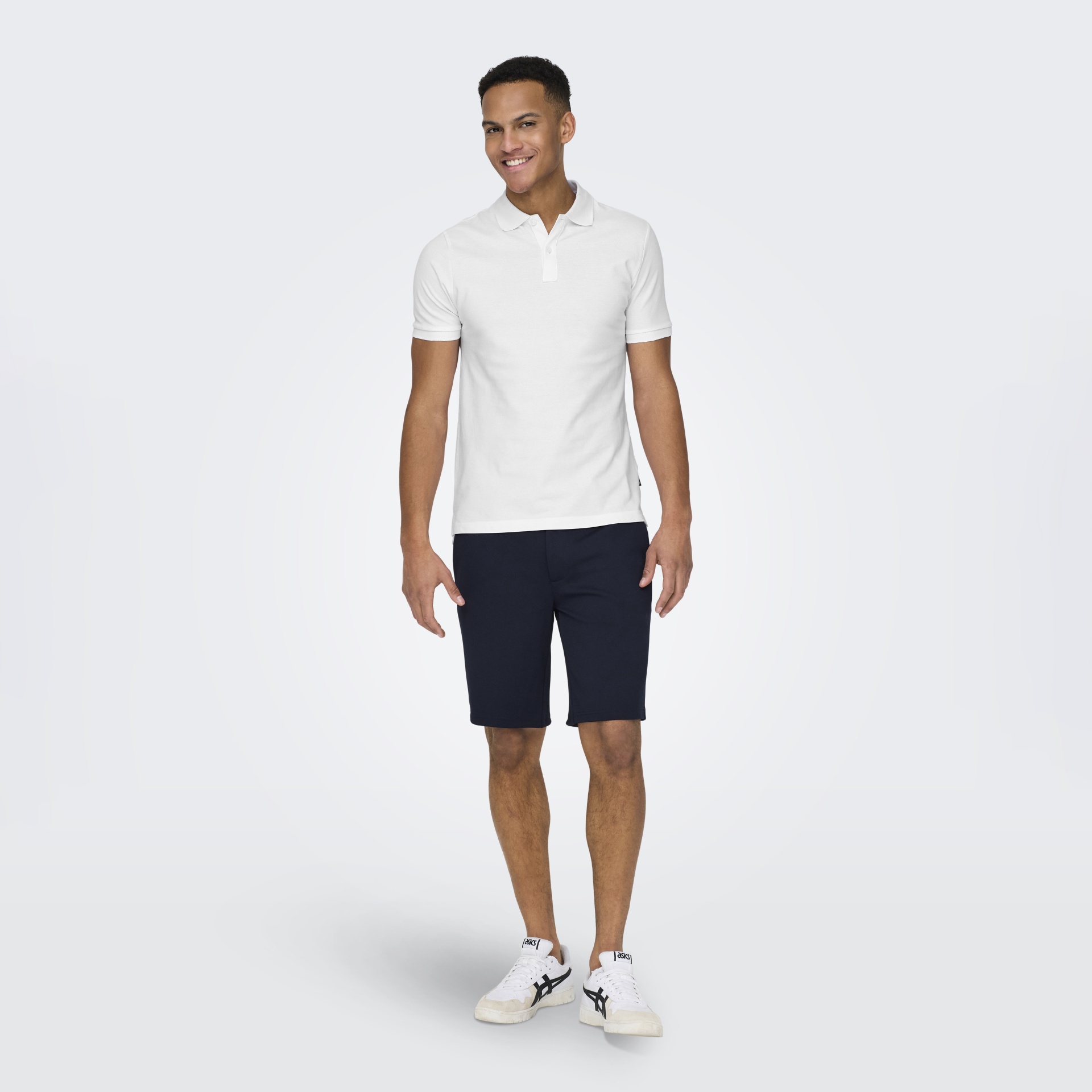 Homem vestido com polo branca, calções pretos e ténis ASICS brancos em fundo branco.