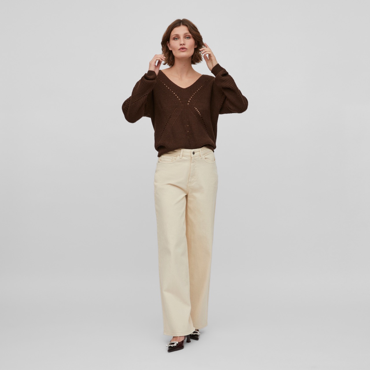 Mulher com camisola castanha de malha e calças largas bege num fundo cinzento claro