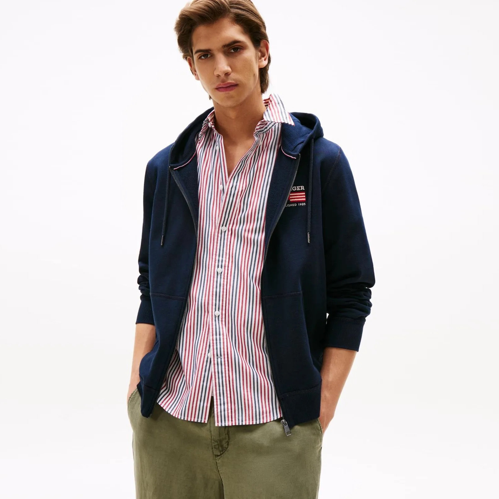 Homem com camisa riscada, casaco azul escuro com capuz e calças verdes.