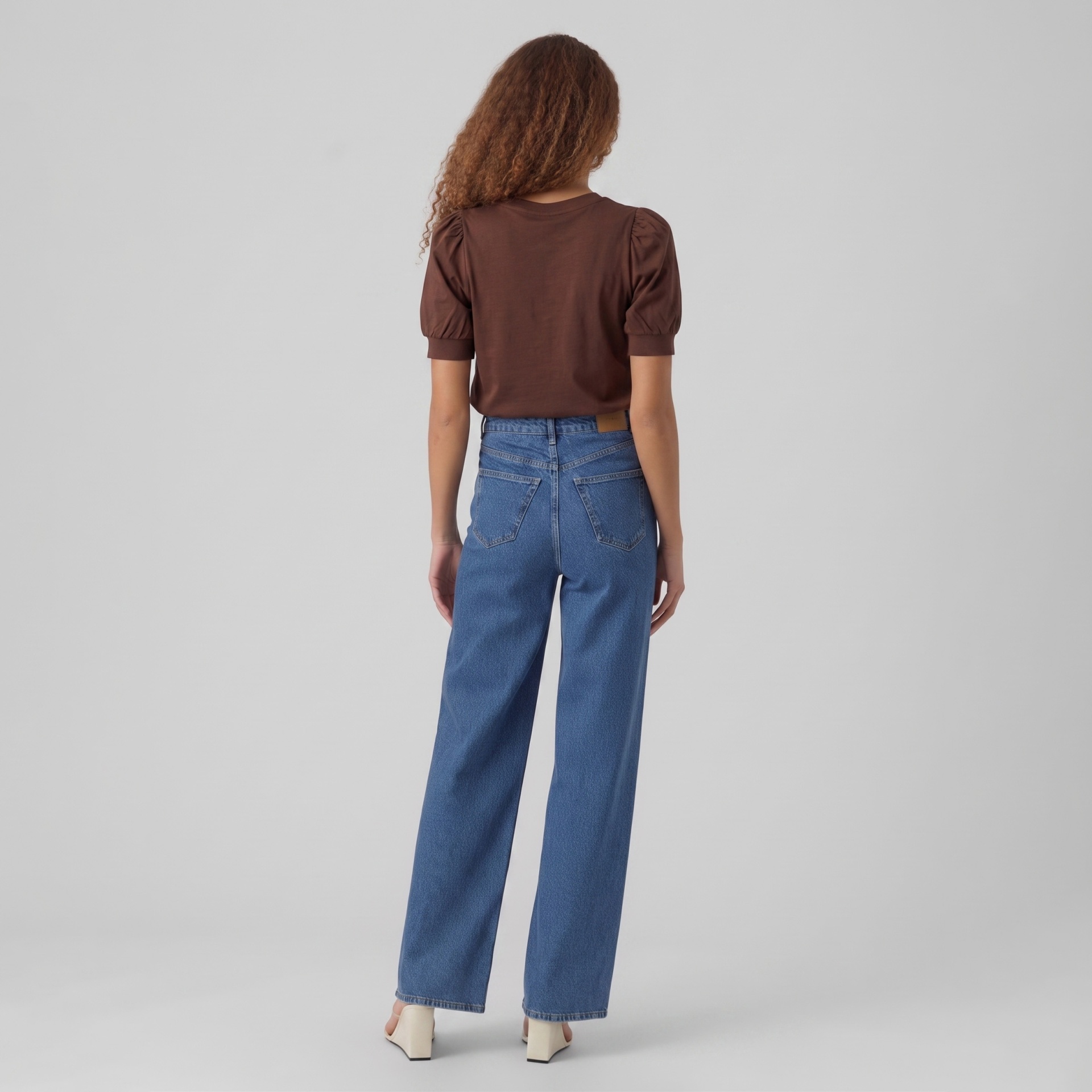 Pessoa de costas com t-shirt castanha, jeans azuis e sapatos brancos de salto.