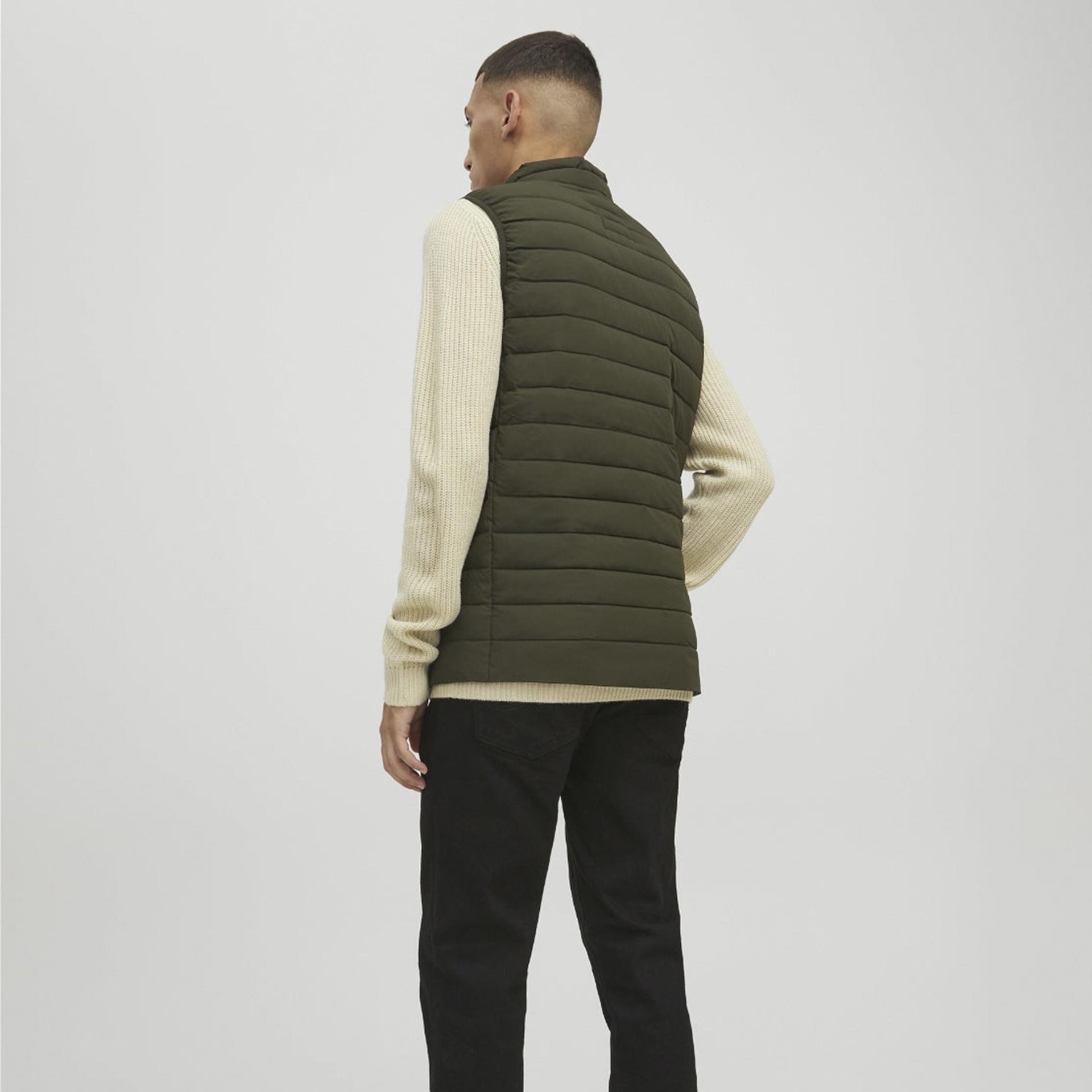 Homem de costas com colete verde-escuro, camisola creme e calças pretas