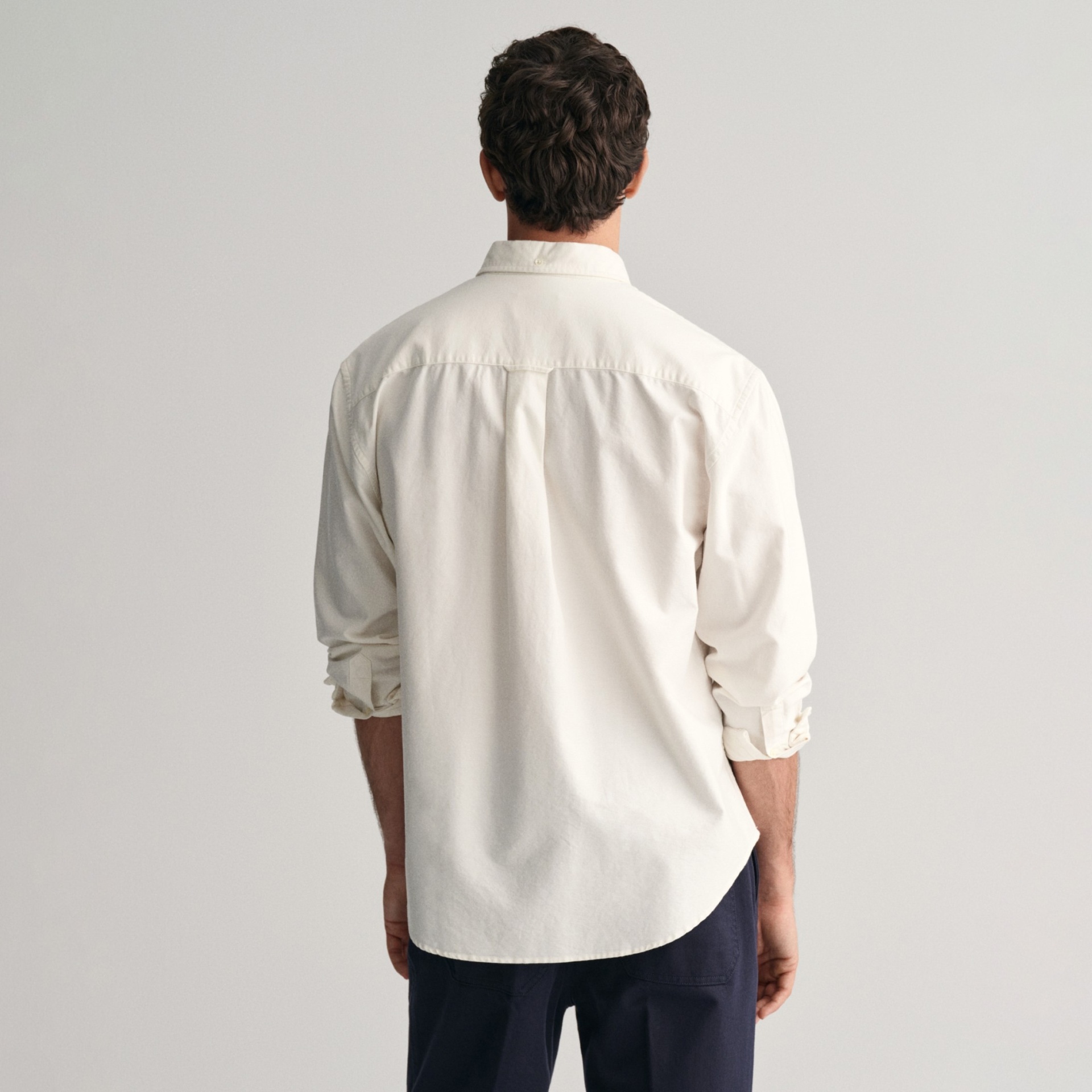 Camisa de algodão creme vista de costas com mangas arregaçadas e calças azul-escuras.