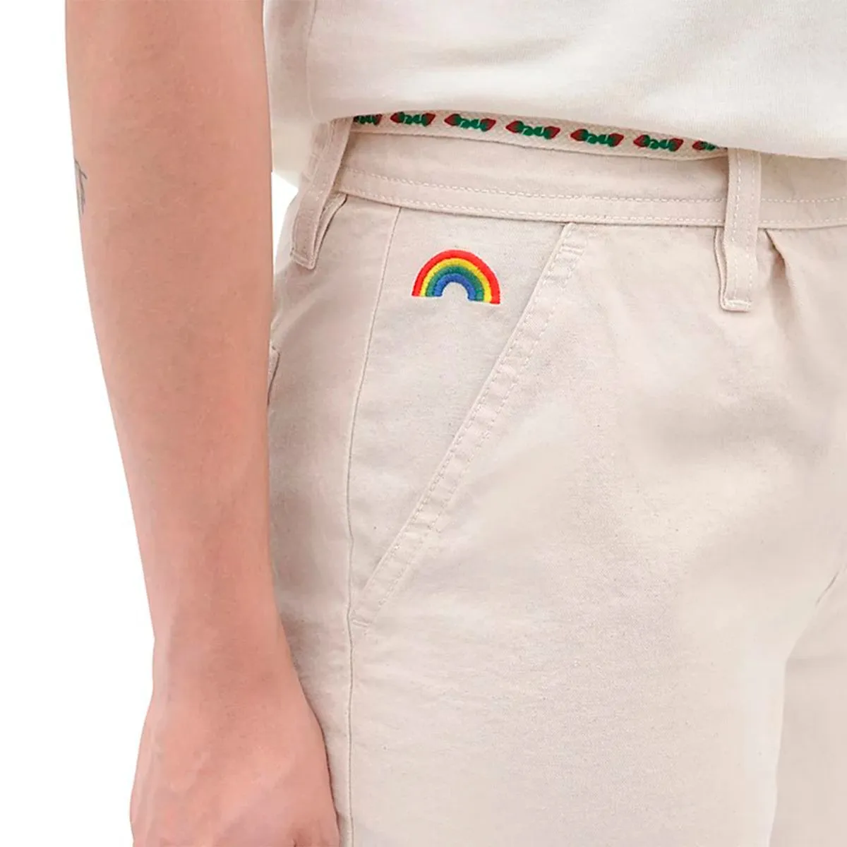 Calças bege com bordado de arco-íris colorido junto ao bolso e cinto com padrão verde e vermelho.