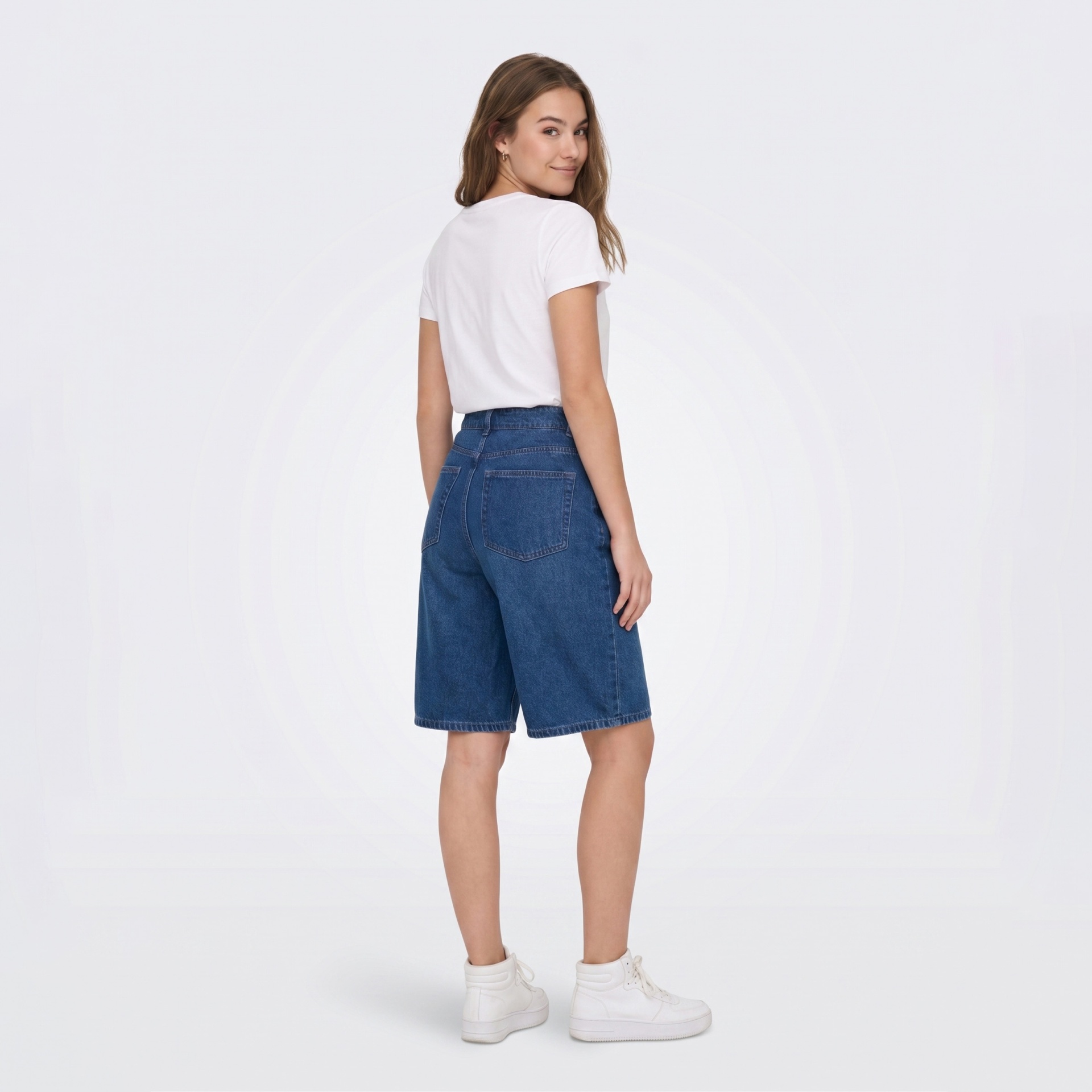 Mulher de costas com t-shirt branca, calções de ganga azuis e sapatilhas brancas num fundo branco.
