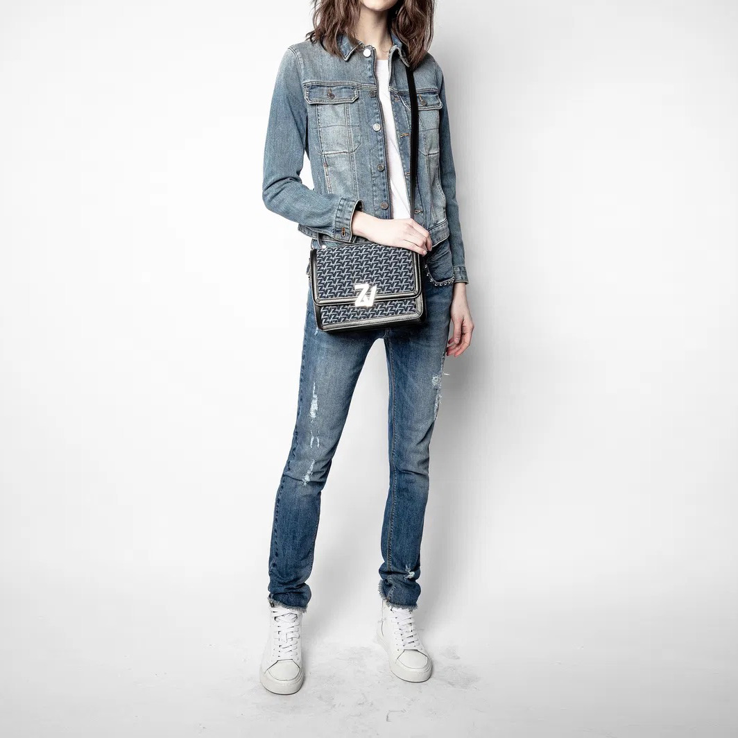 Mulher com casaco e jeans de ganga azul e ténis brancos, segurando mala preta com padrão e fecho prateado