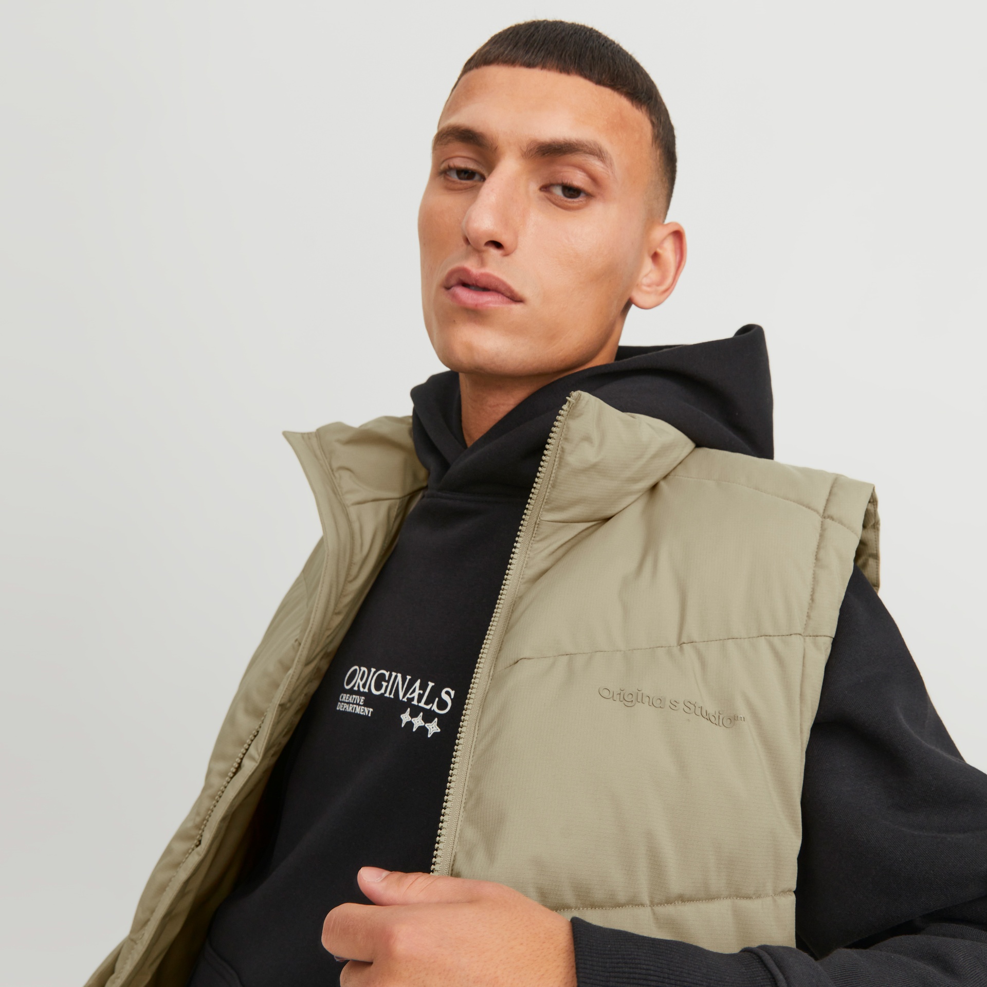 Homem com colete acolchoado bege claro por cima de hoodie preto com texto branco numa parede cinza claro