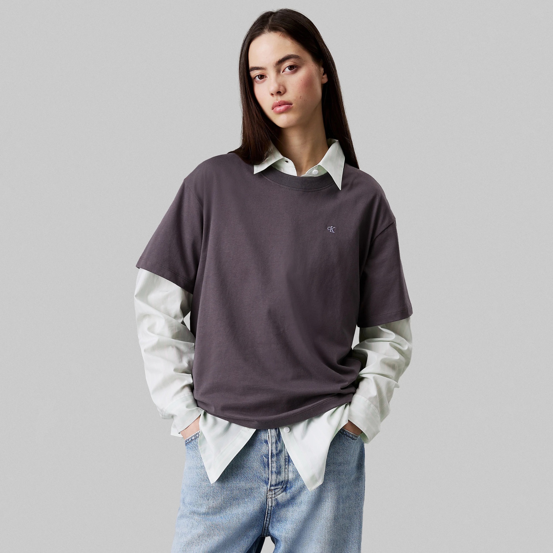 Mulher com camisola cinzenta e camisa branca de manga comprida com calças de ganga azuis claras