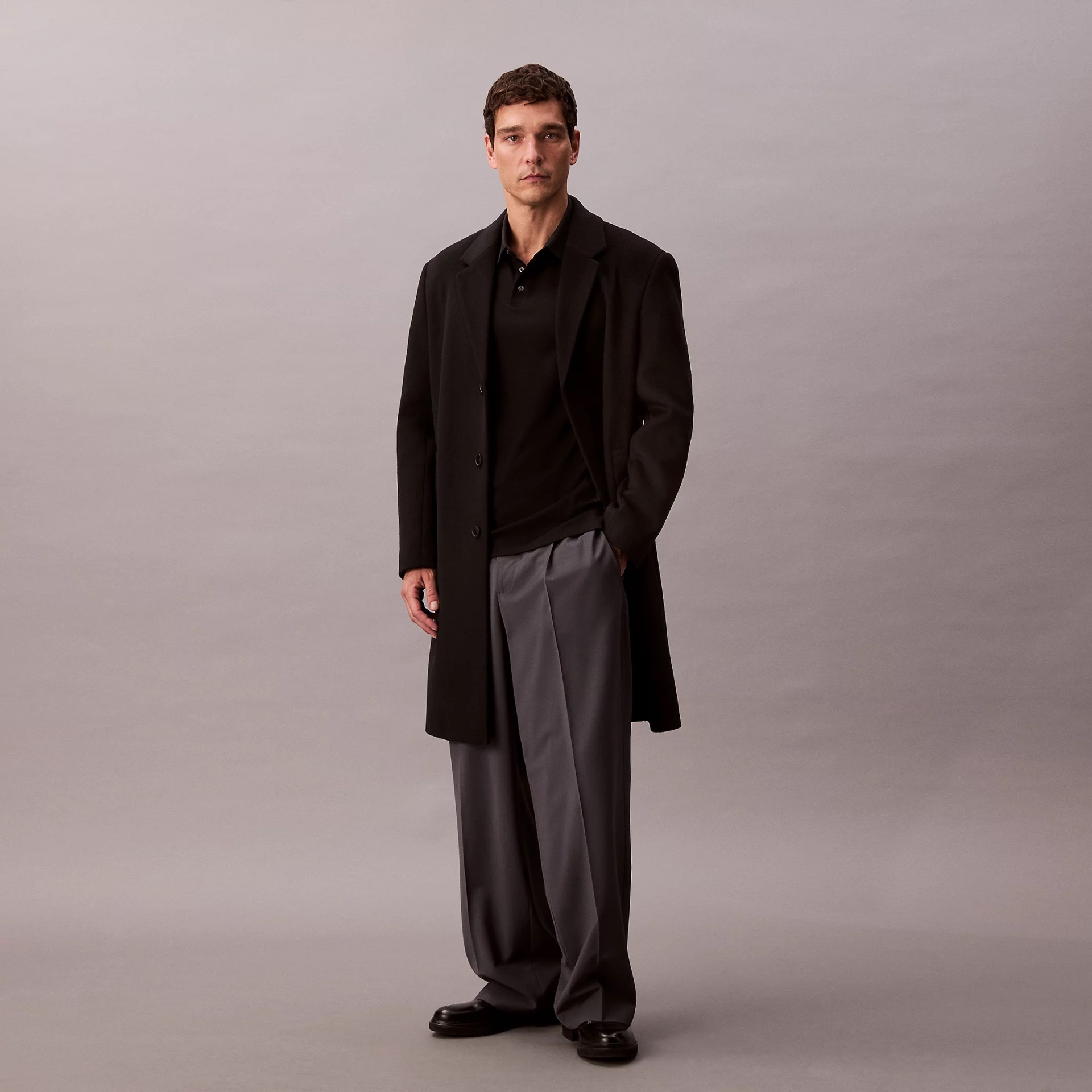Homem com casaco preto, camisa polo preta e calças cinzentas