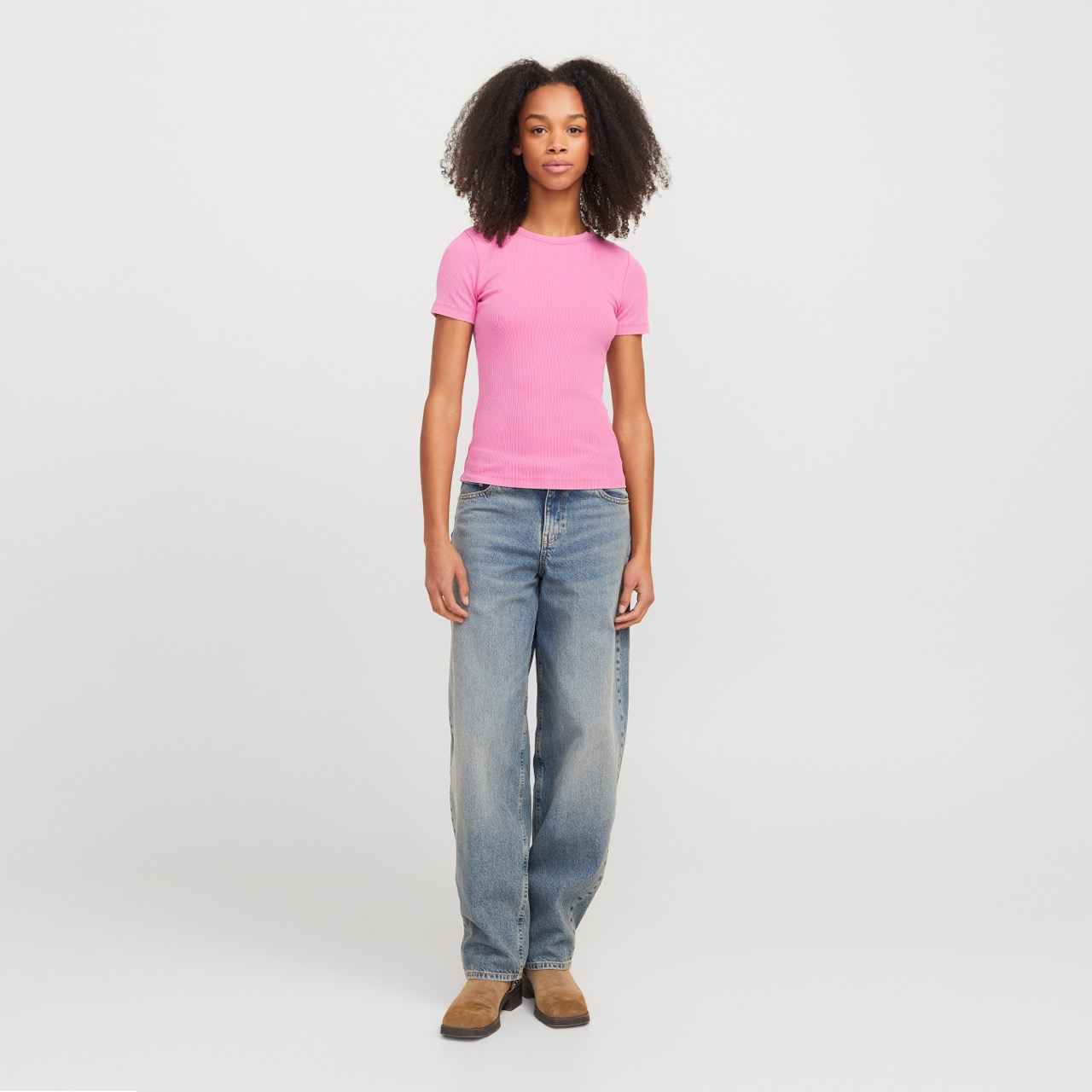Mulher a usar t-shirt rosa canelada, jeans azuis e sapatos marrons, em fundo branco