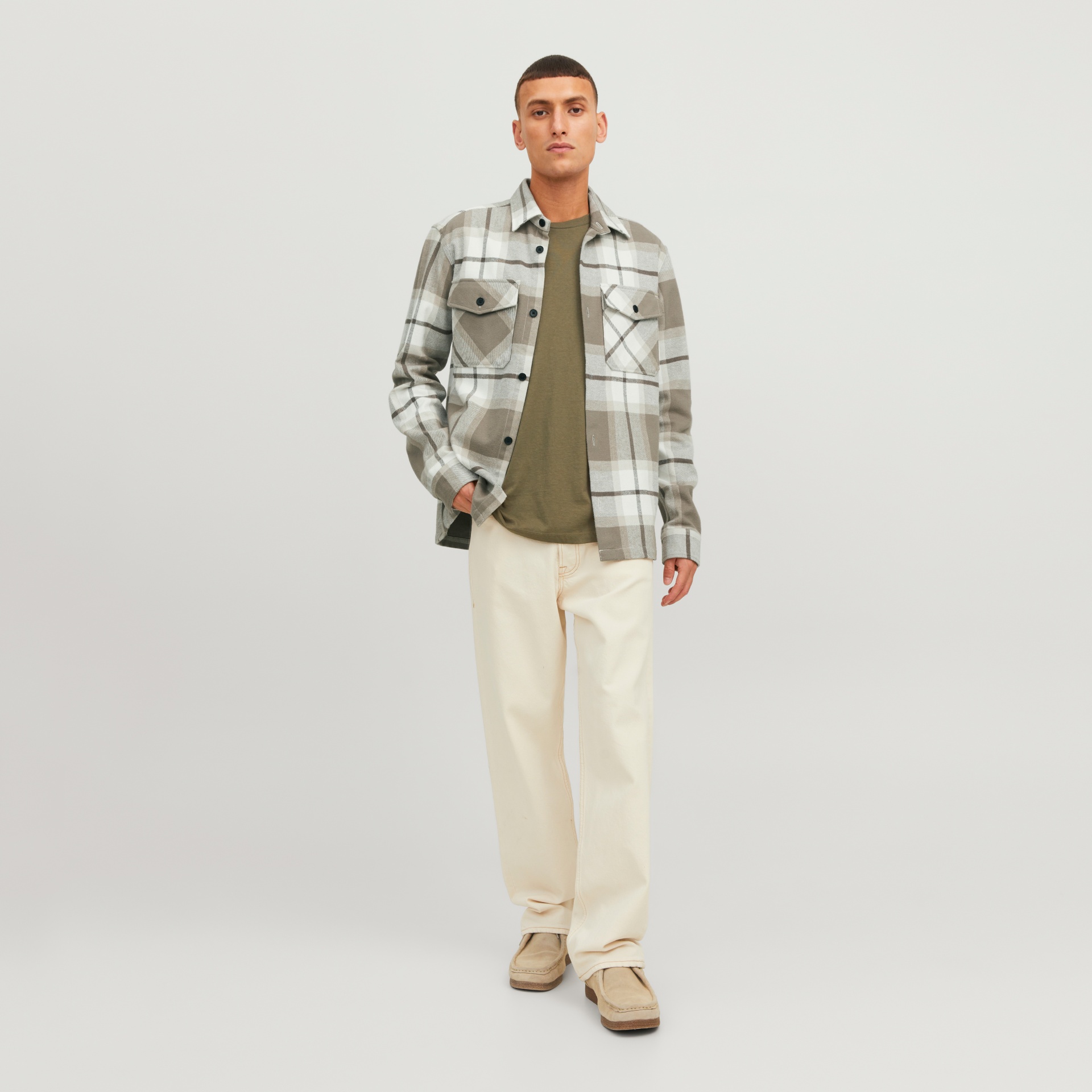 Homem com camisa xadrez bege, branco e castanho, t-shirt verde, calças creme largas e sapatos castanhos claros em fundo branco