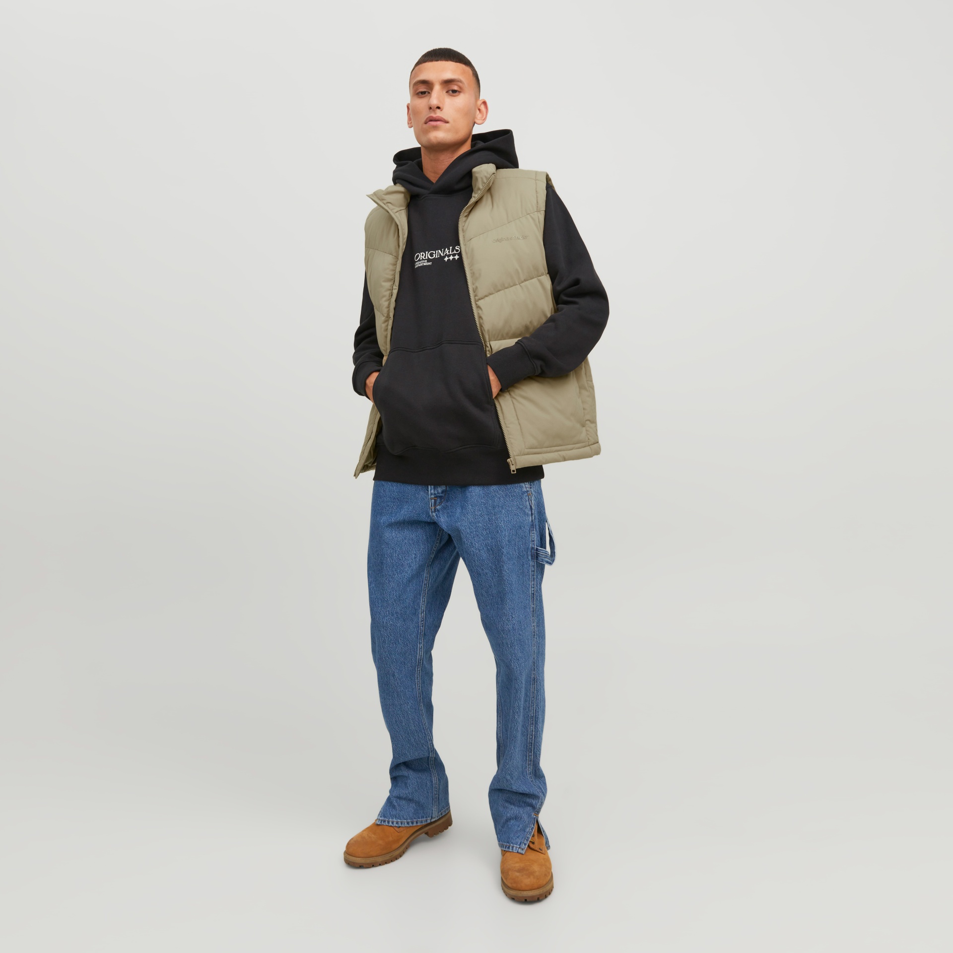 Homem com colete bege acolchoado, sweat preto com capuz e texto, jeans azul e sapatos castanhos