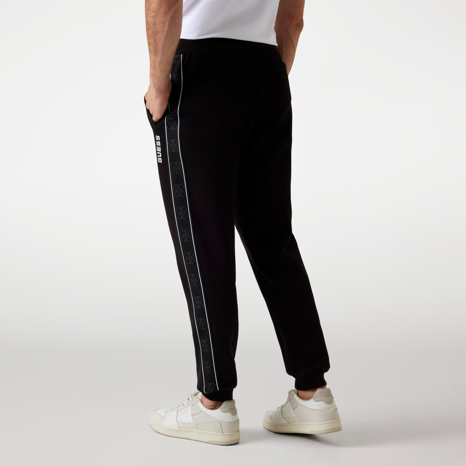 Calças de treino pretas com faixa lateral branca e texto GUESS, usadas com sapatilhas brancas.