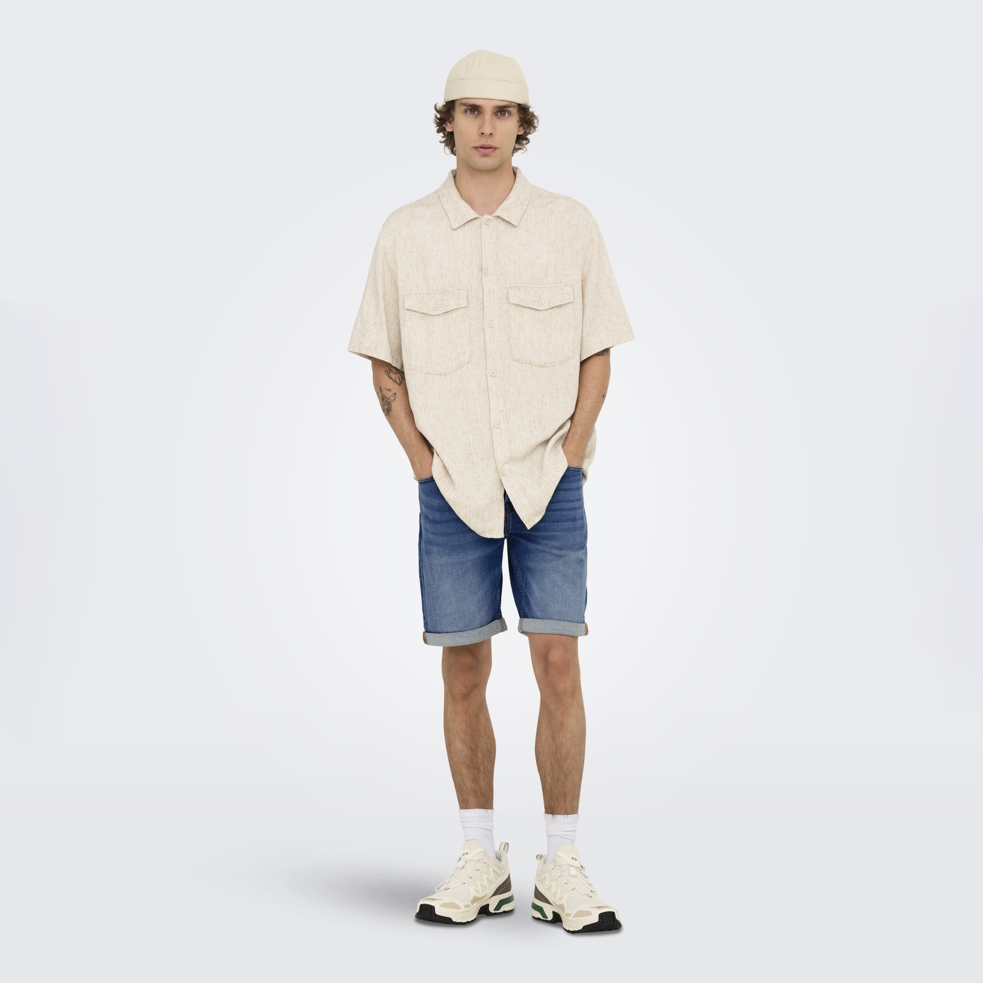 Homem a usar camisa bege, boné bege, calções de ganga e ténis brancos.
