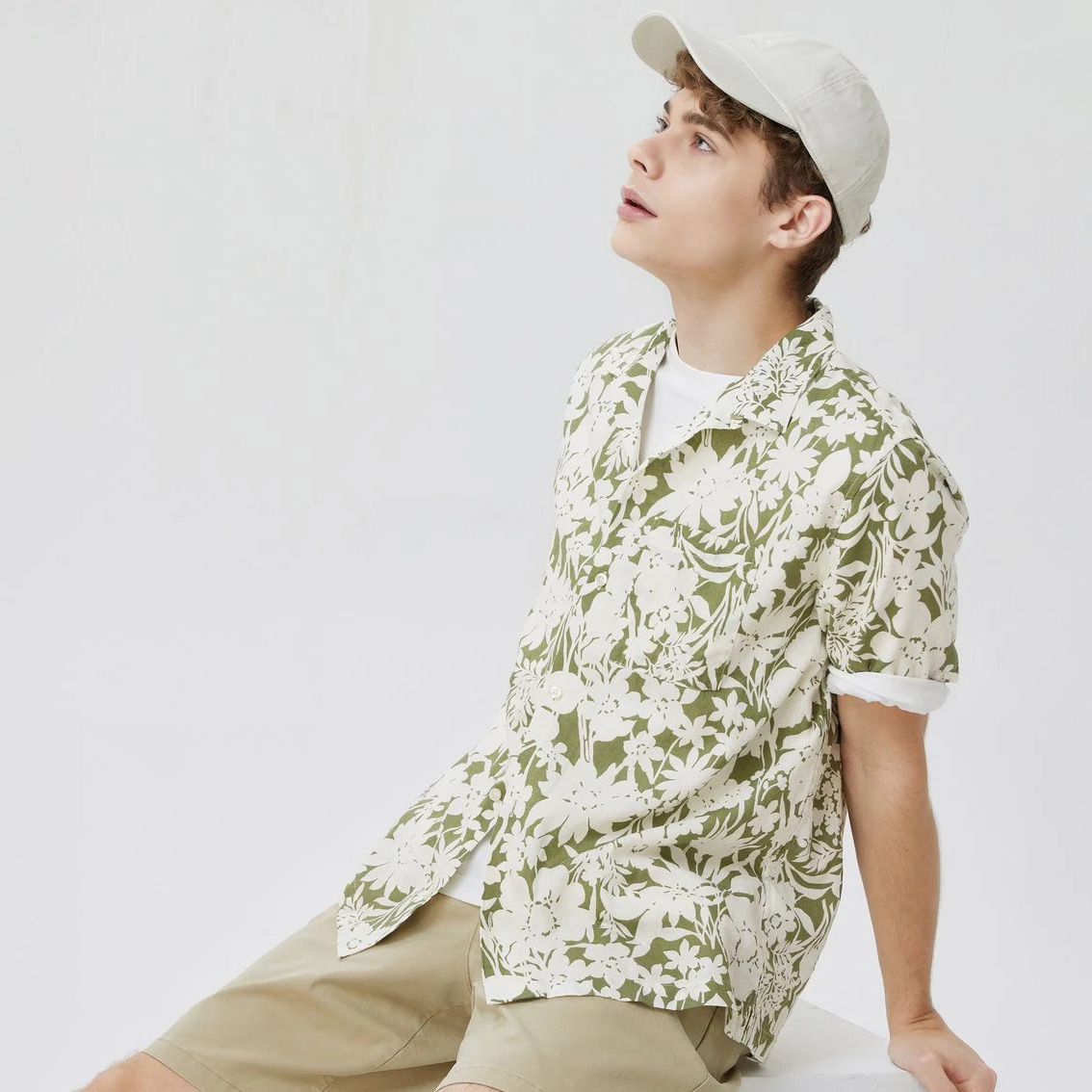 Homem com camisa verde floral, t-shirt branca, calções e boné bege sentado.