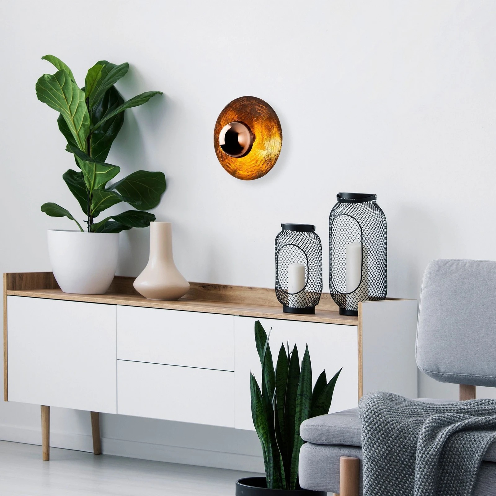 Sala minimalista com móvel branco e madeira, plantas verdes, aplique dourado e cadeira cinzenta.