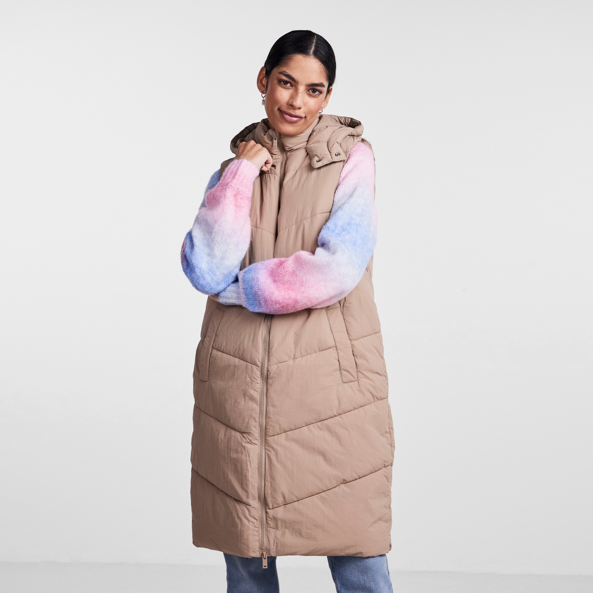 Colete comprido bege acolchoado com capuz e zíper, usado por mulher com camisola colorida de malha