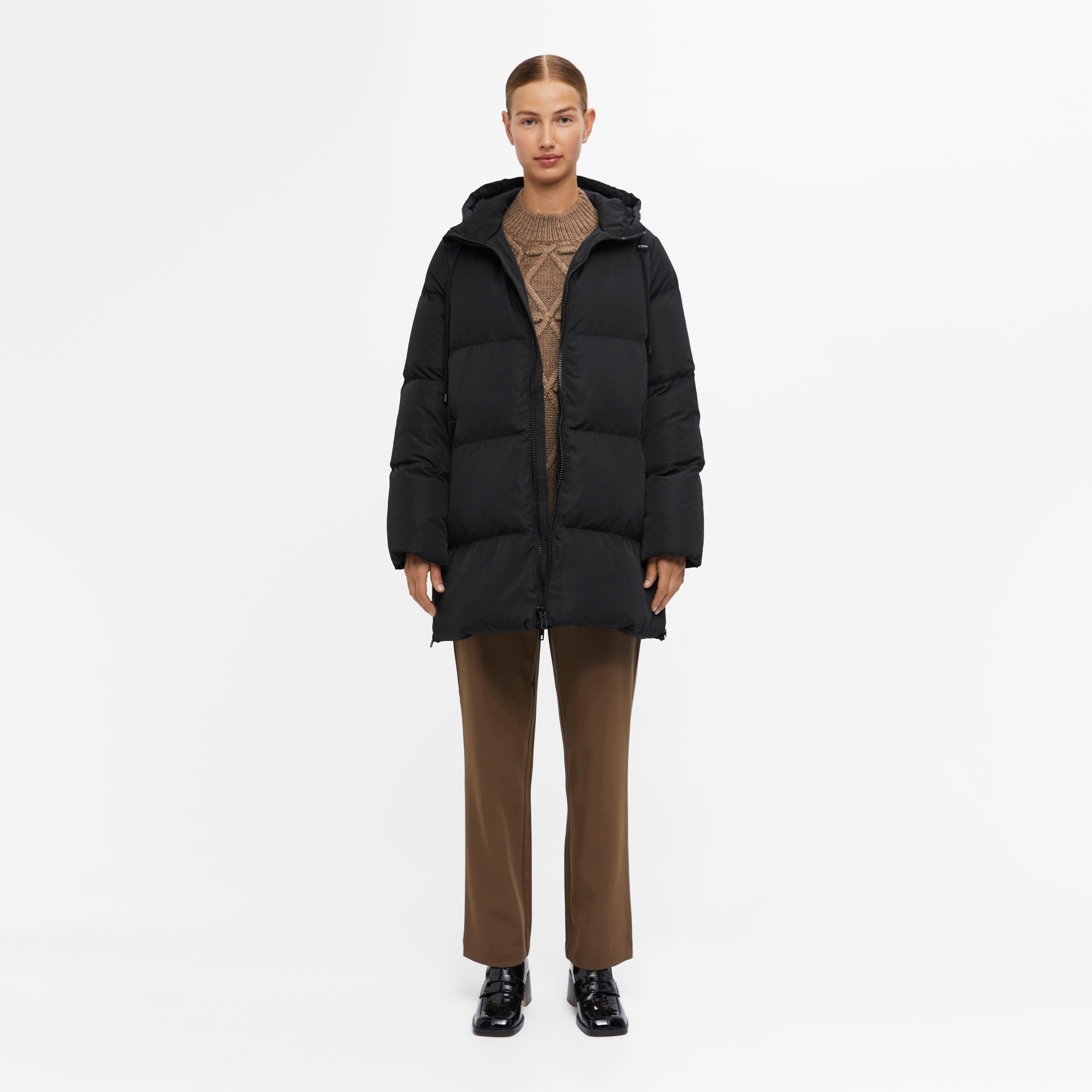 Mulher com casaco acolchoado preto e capuz, camisola castanha, calças castanhas e sapatos pretos em fundo branco.