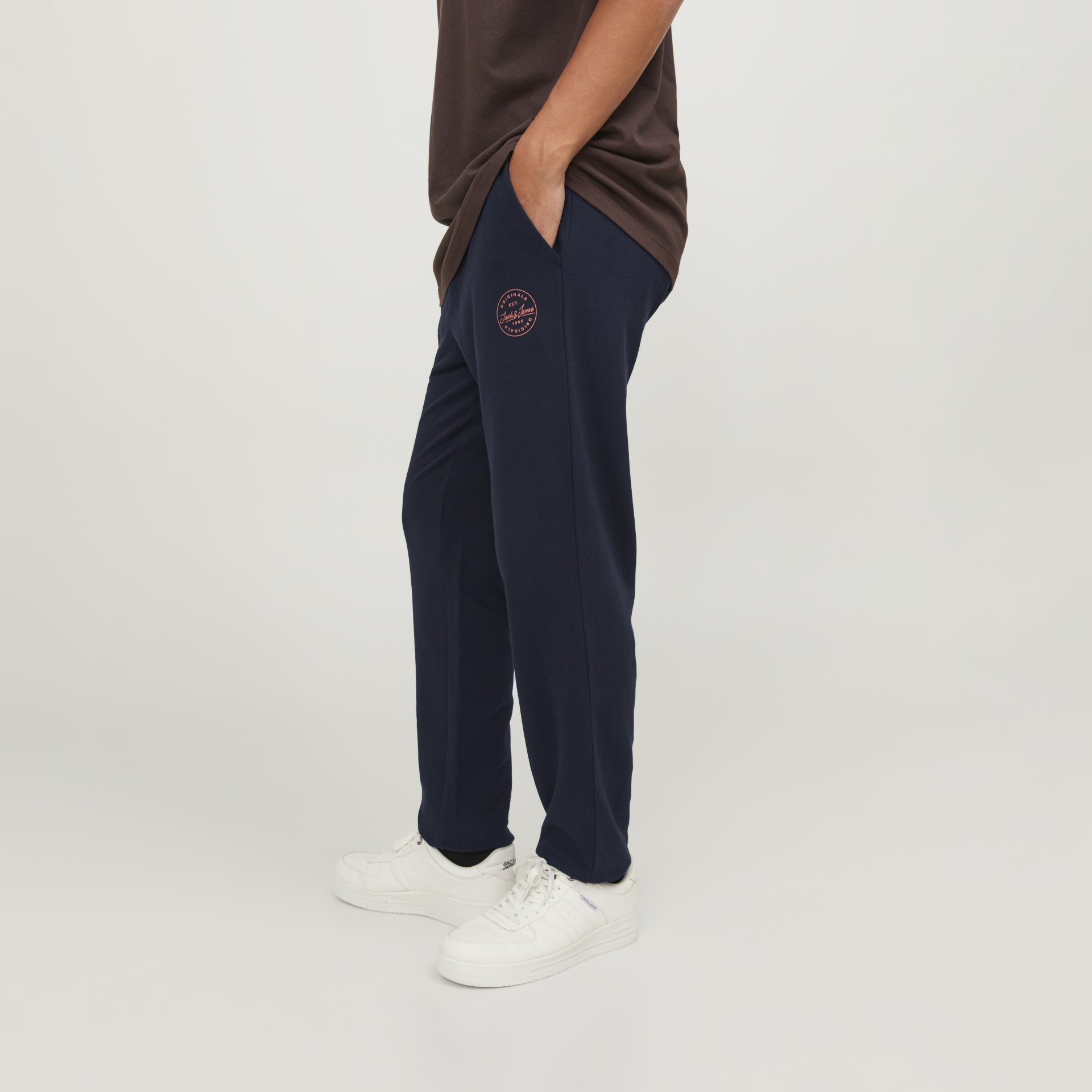 Calças de tecido azul escuro com logótipo rosa e bolso lateral, usadas com t-shirt castanha e ténis brancos.