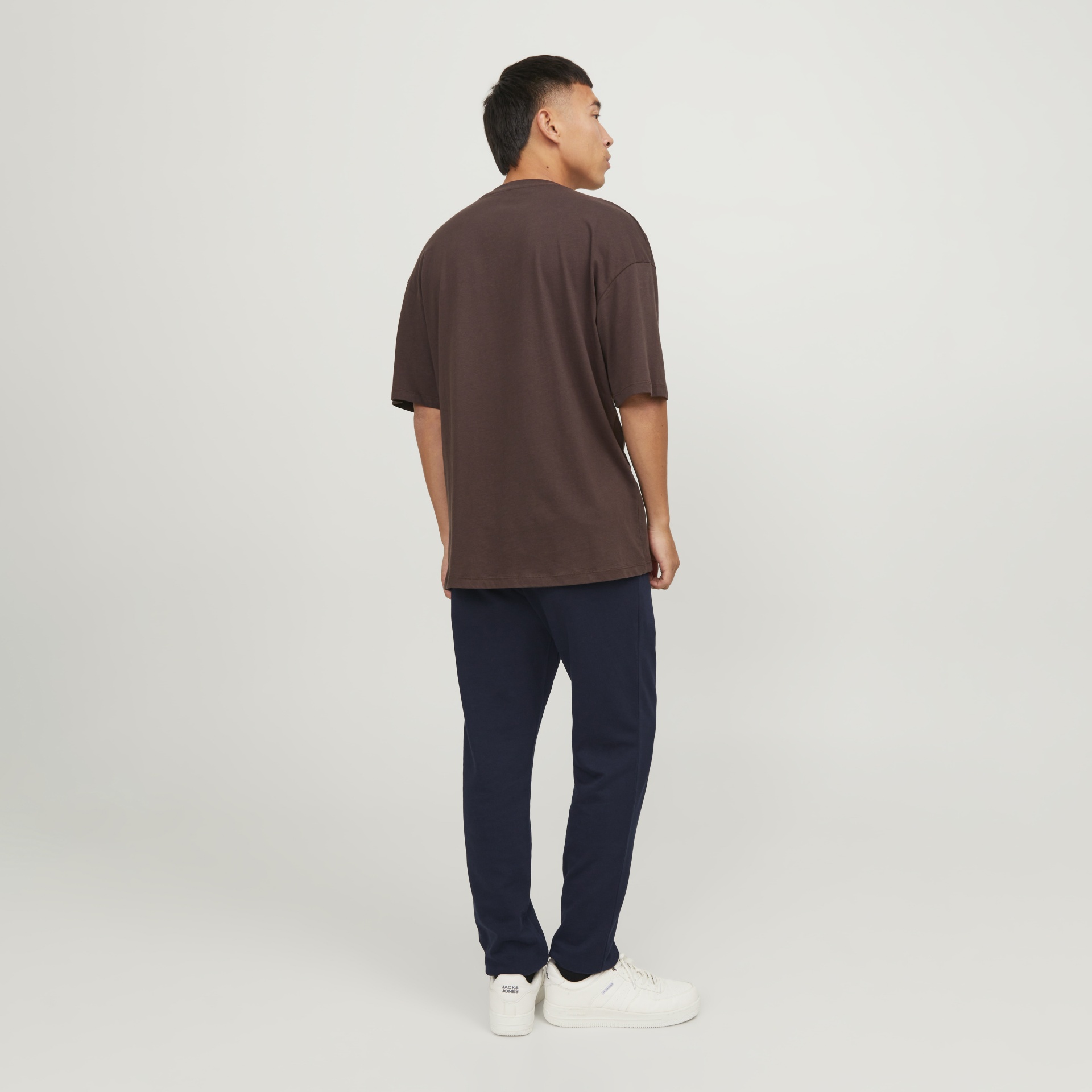 Homem de costas com t-shirt castanha, calças azuis e sapatilhas brancas JACK & JONES