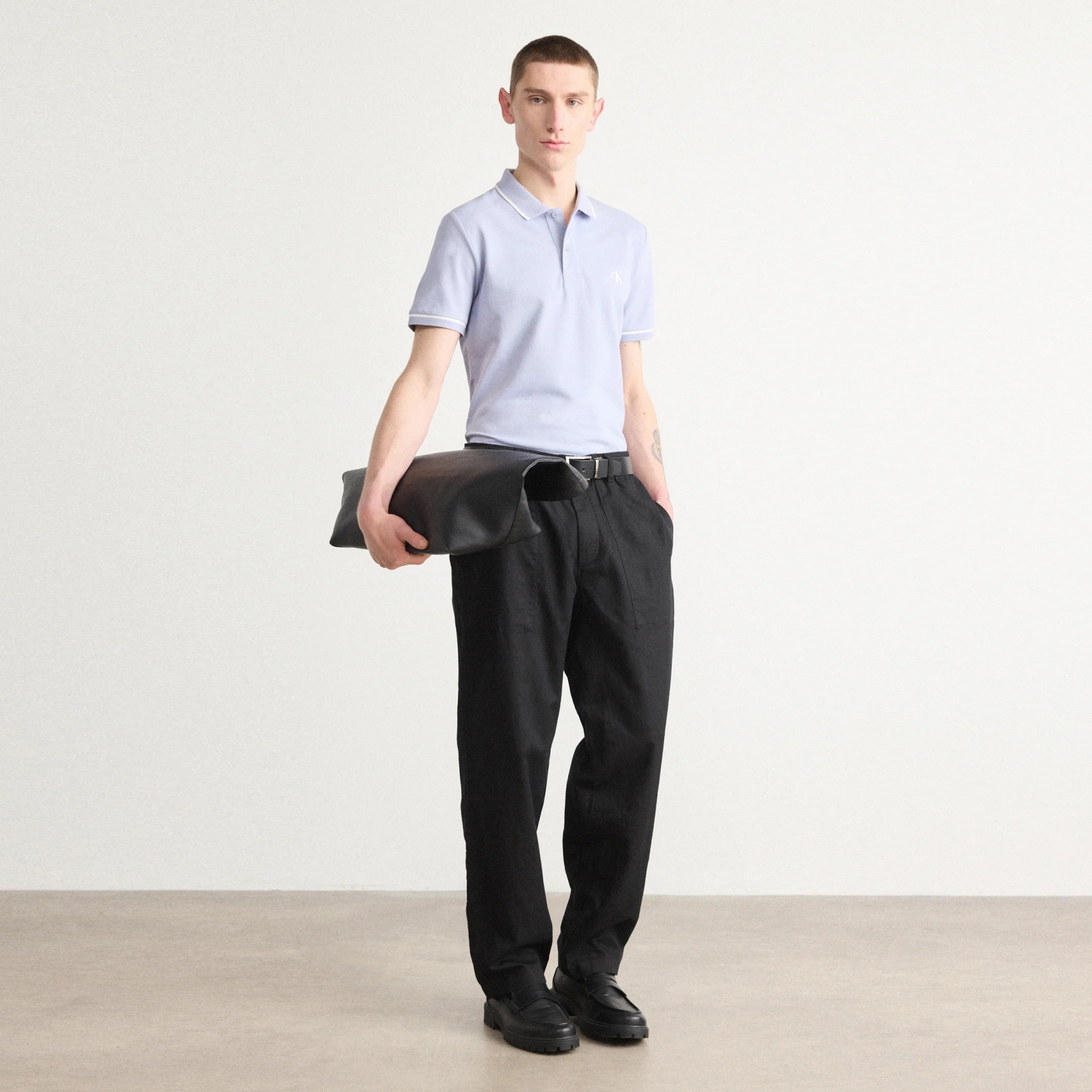 Homem com polo azul claro, calças pretas, cinto e sapatos pretos segurando bolsa de couro preta