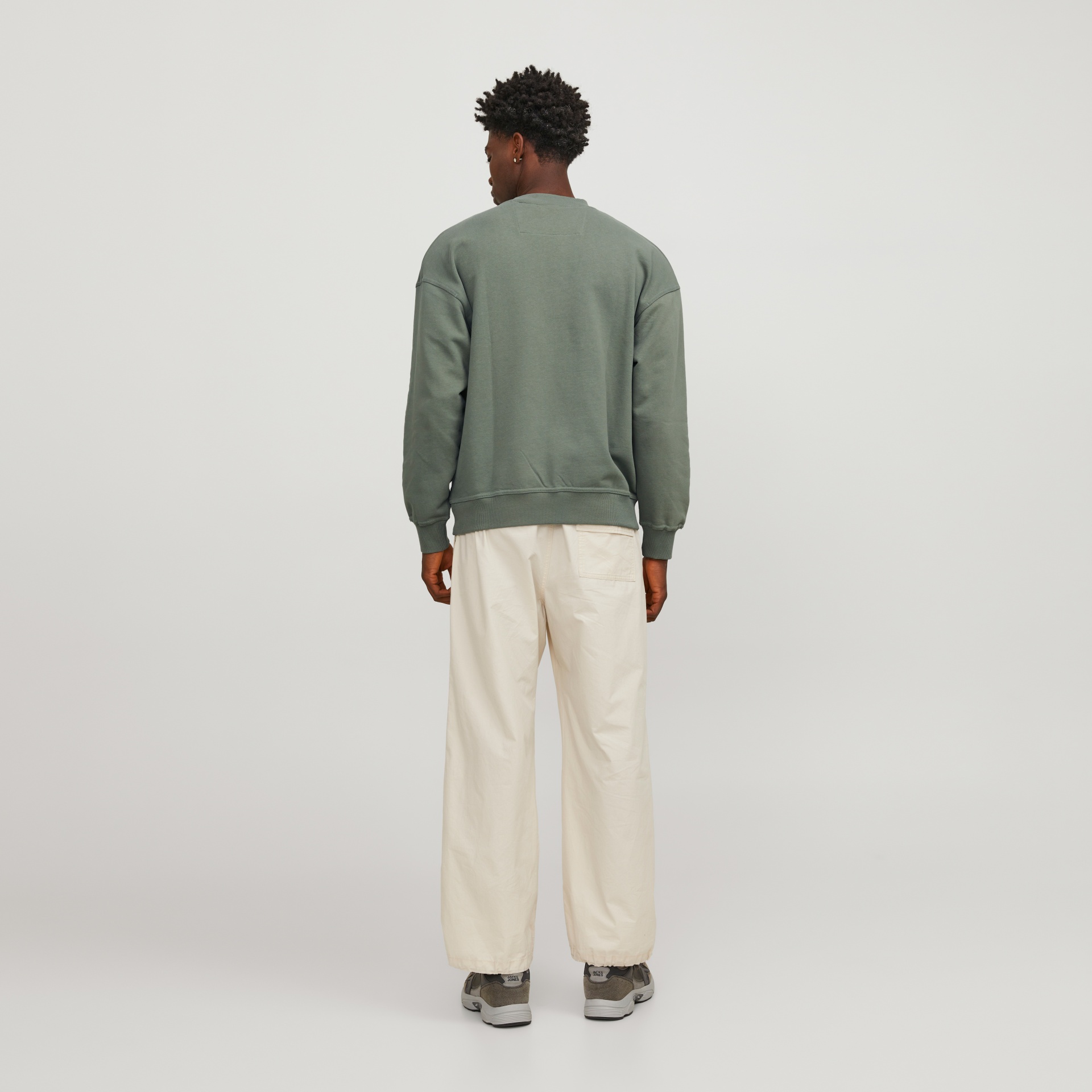 Homem de costas com sweat cinza esverdeado, calças brancas largas e ténis cinzentos em fundo branco.