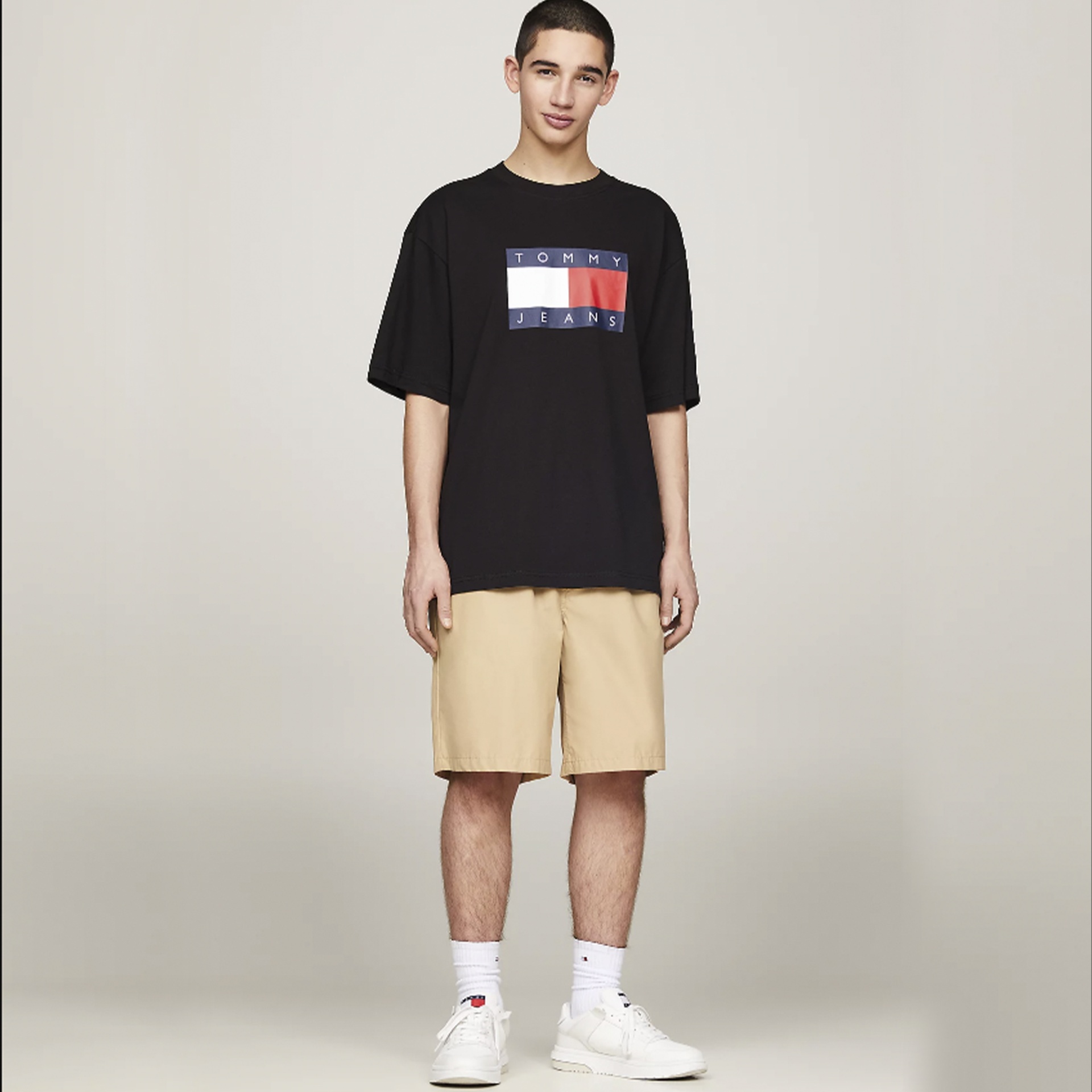 Homem com t-shirt preta Tommy Jeans, calções bege, meias e sapatilhas brancas em fundo cinzento claro