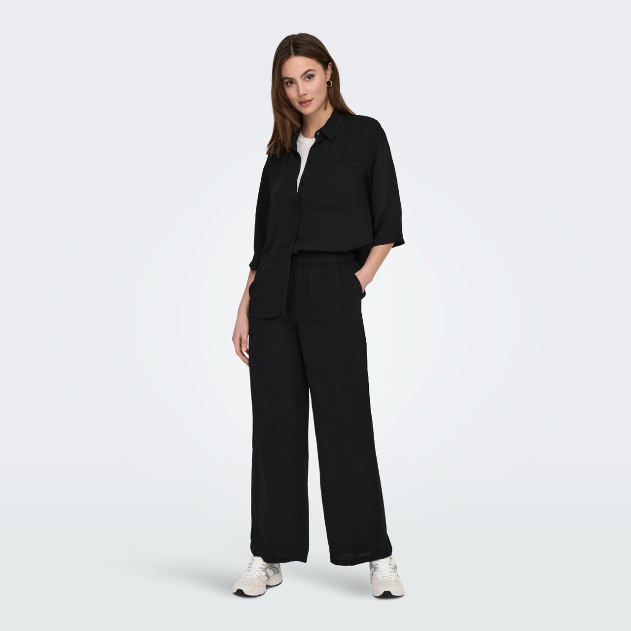 Mulher com conjunto preto de camisa e calças largas com sapatilhas brancas, fundo branco.