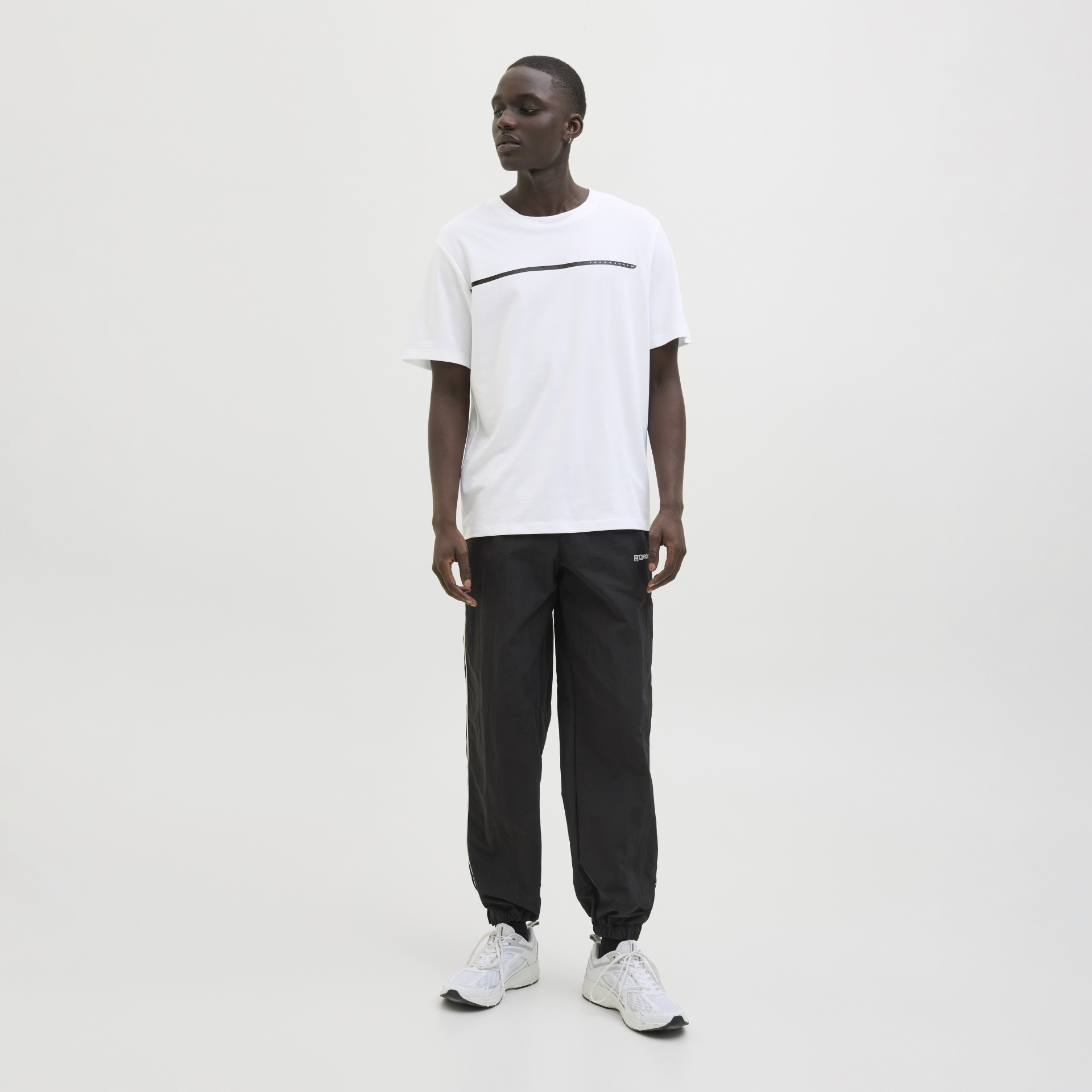 Homem com t-shirt branca, calças pretas largas e ténis brancos num fundo branco