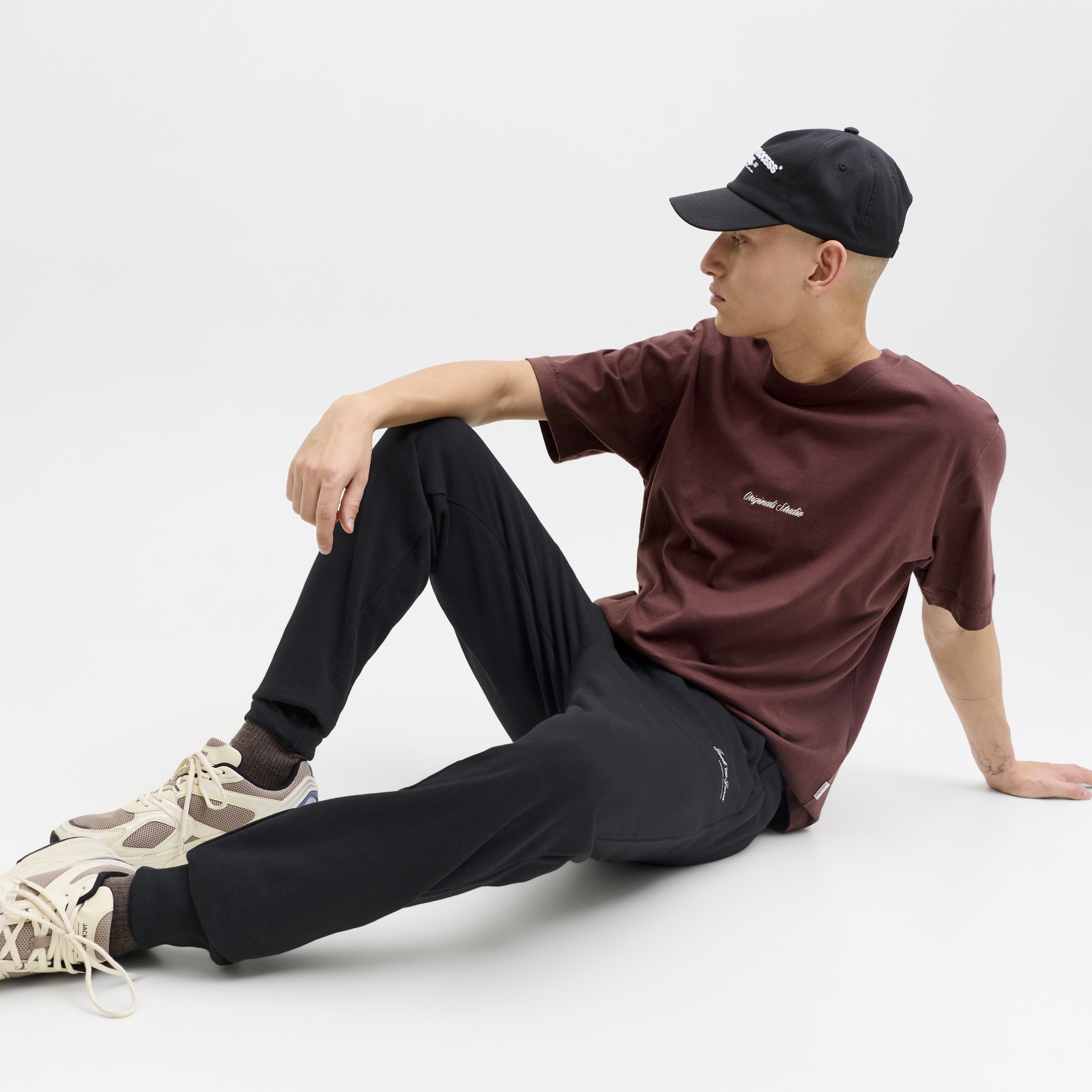 Homem com roupa casual e boné preto sentado num fundo branco.