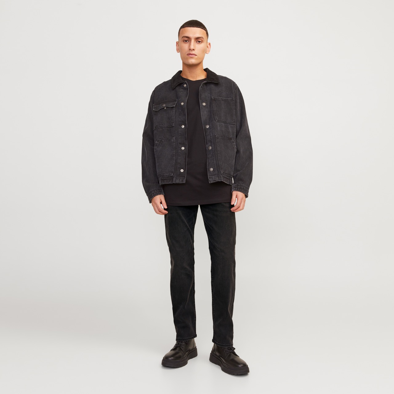 Homem com camisola preta de ganga, t-shirt preta, calças pretas e sapatos pretos em fundo branco.