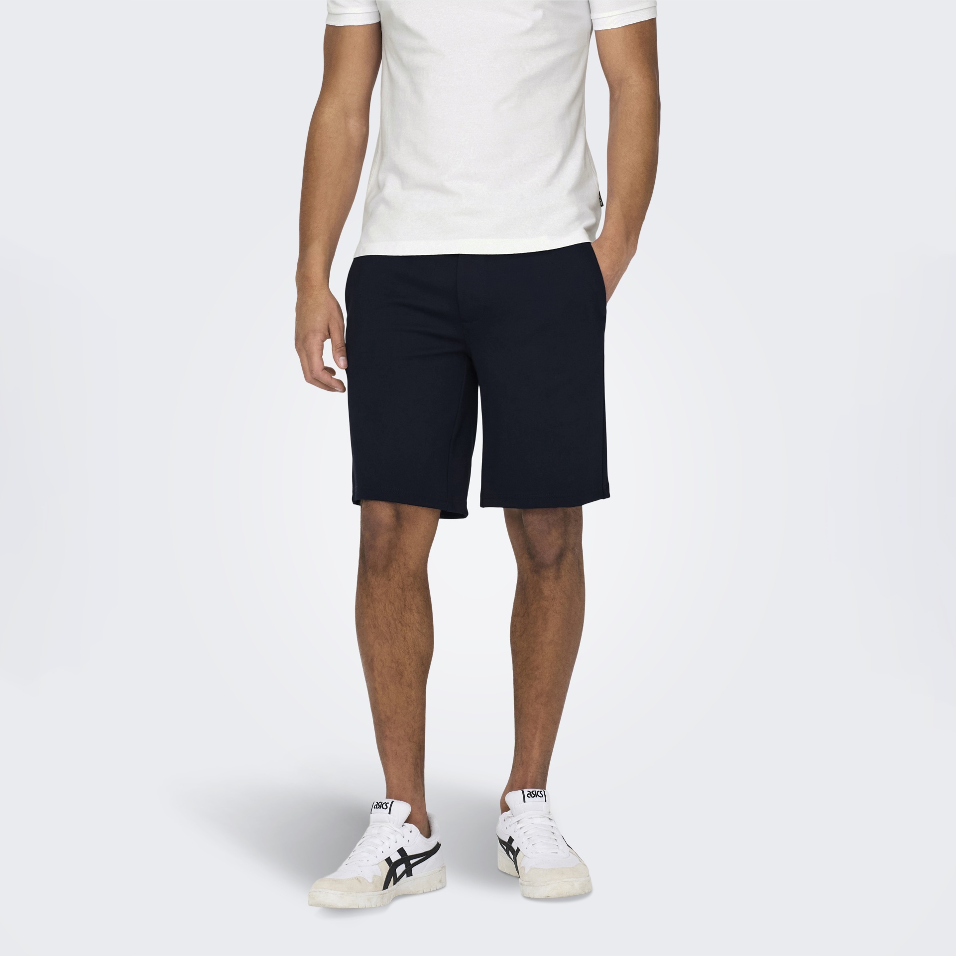 Homem com t-shirt branca, calções pretos e sapatilhas ASICS brancas pretas em fundo cinza