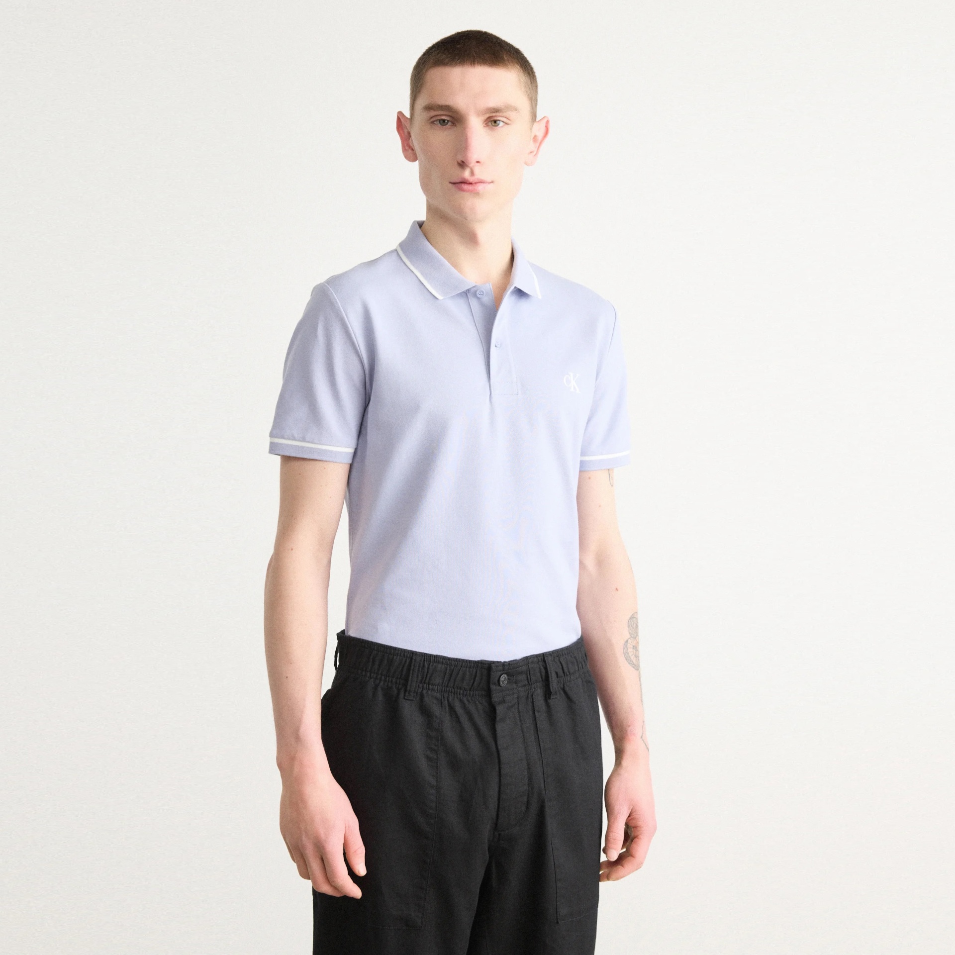 Homem com polo azul claro e calças pretas em fundo branco.