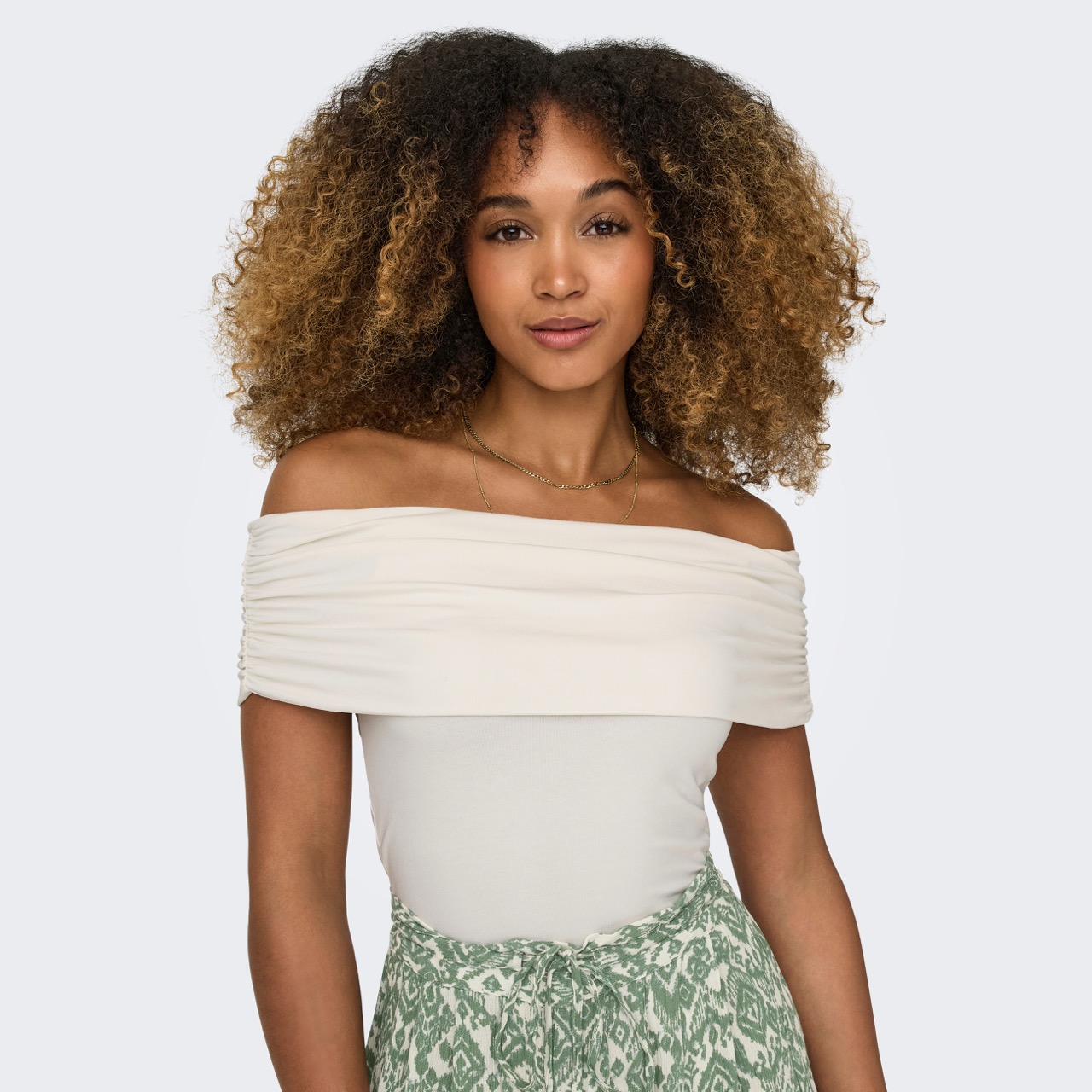 Mulher com blusa branca ombros descobertos e calças verdes com padrão branco